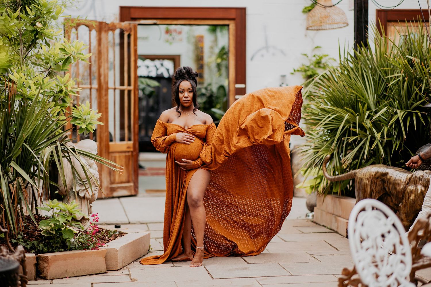 Luxury picture of Pregnant woman in flowing rust orange maternity gown poses outdoors in lush garden courtyard.