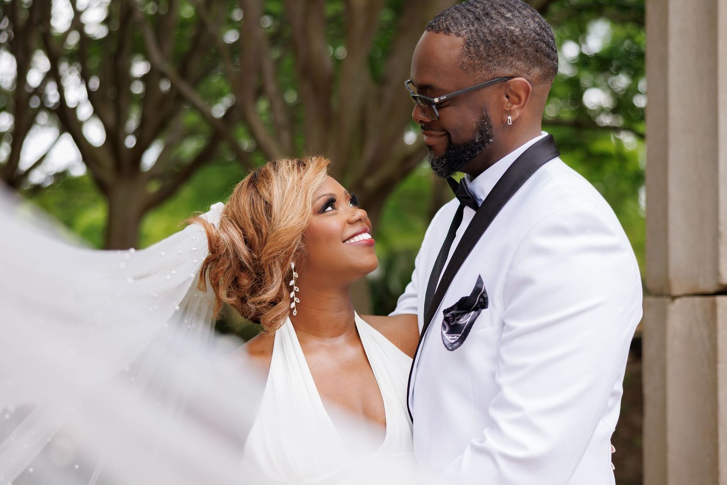 Bride and groom share a romantic moment outdoors, bride in white gown with veil, groom in white suit with black lapels.