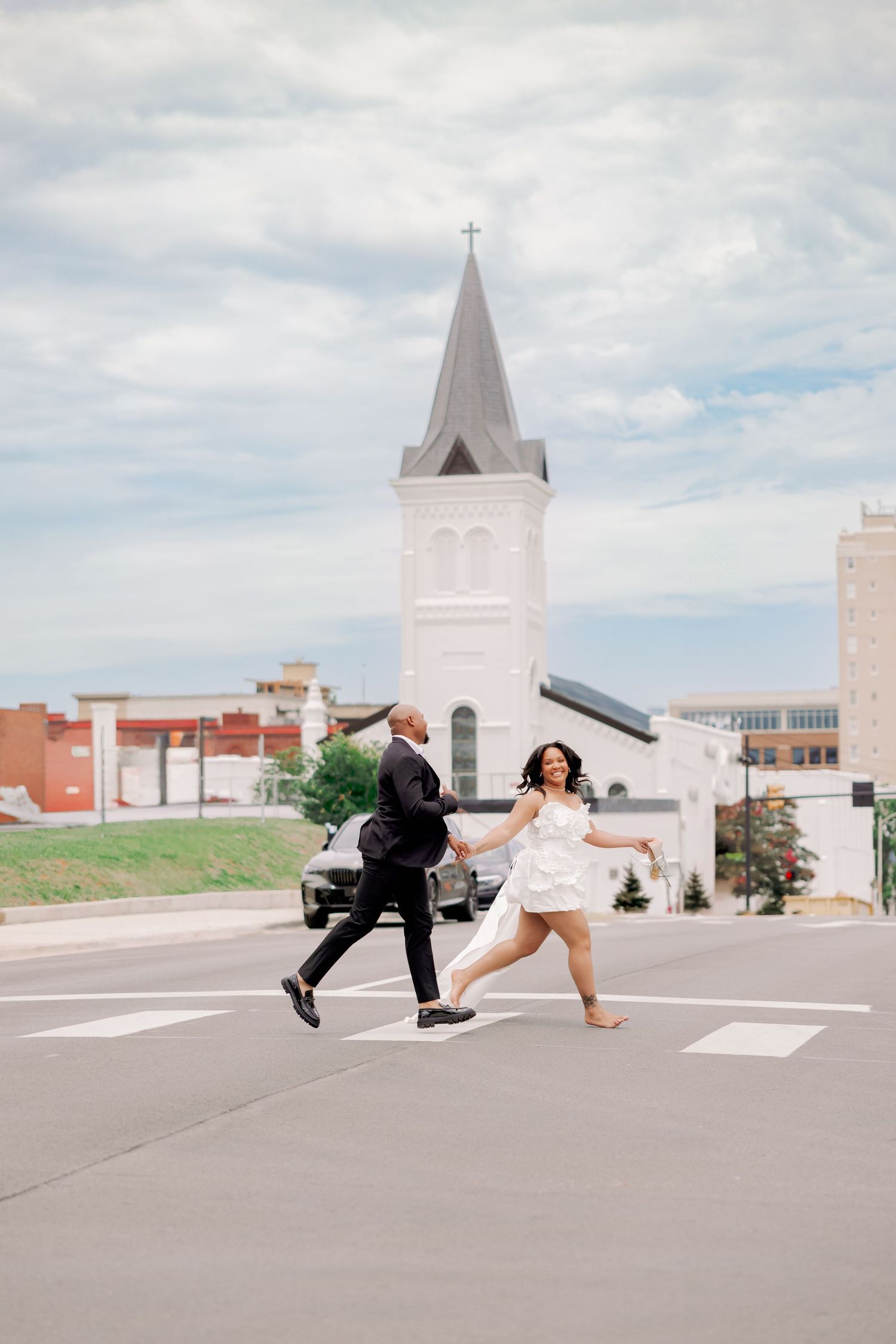 A joyful couple in wedding attire runs across a crosswalk with a white church steeple in the background.