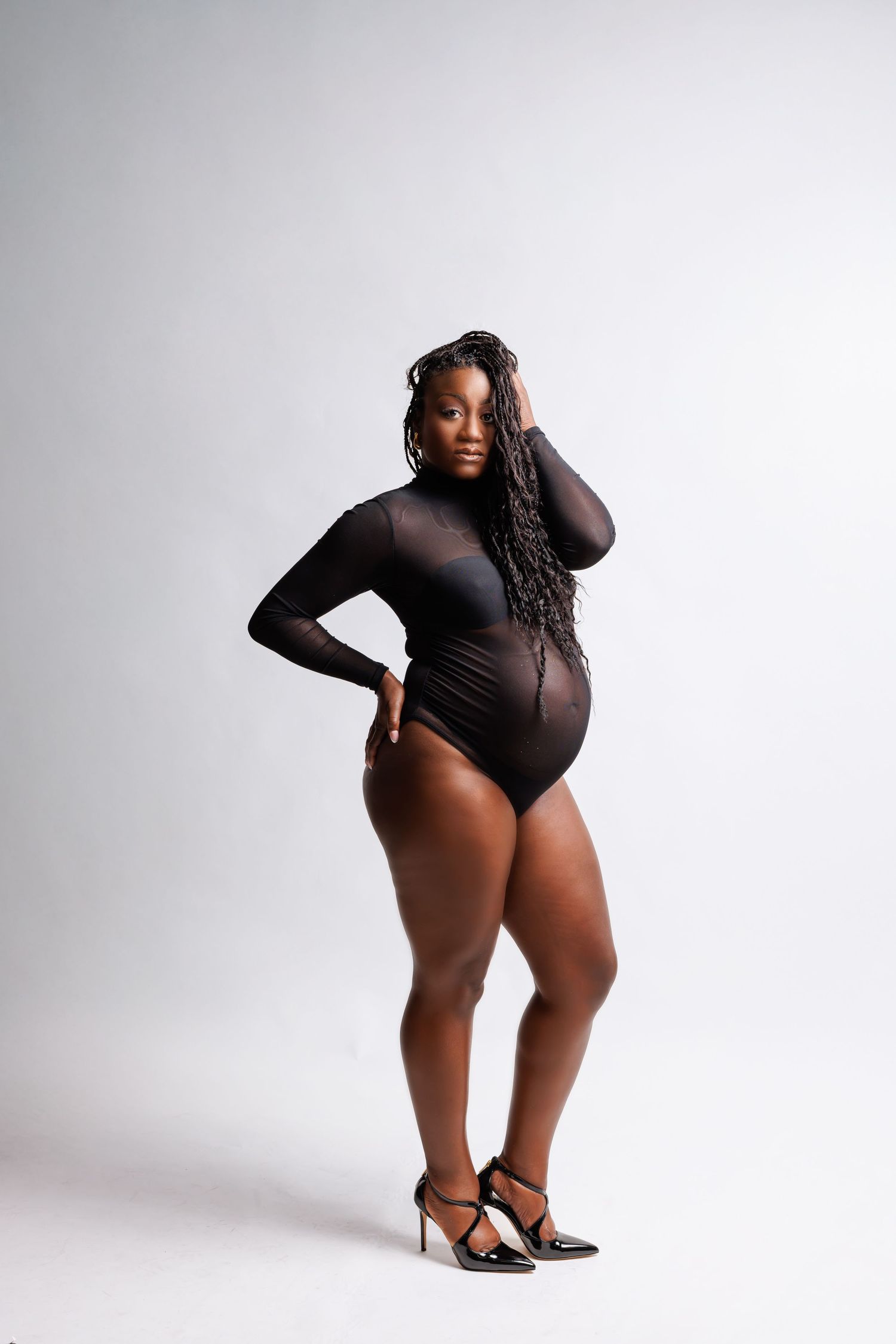Plus-size Black woman in black mesh bodysuit and heels posing confidently against white background.
