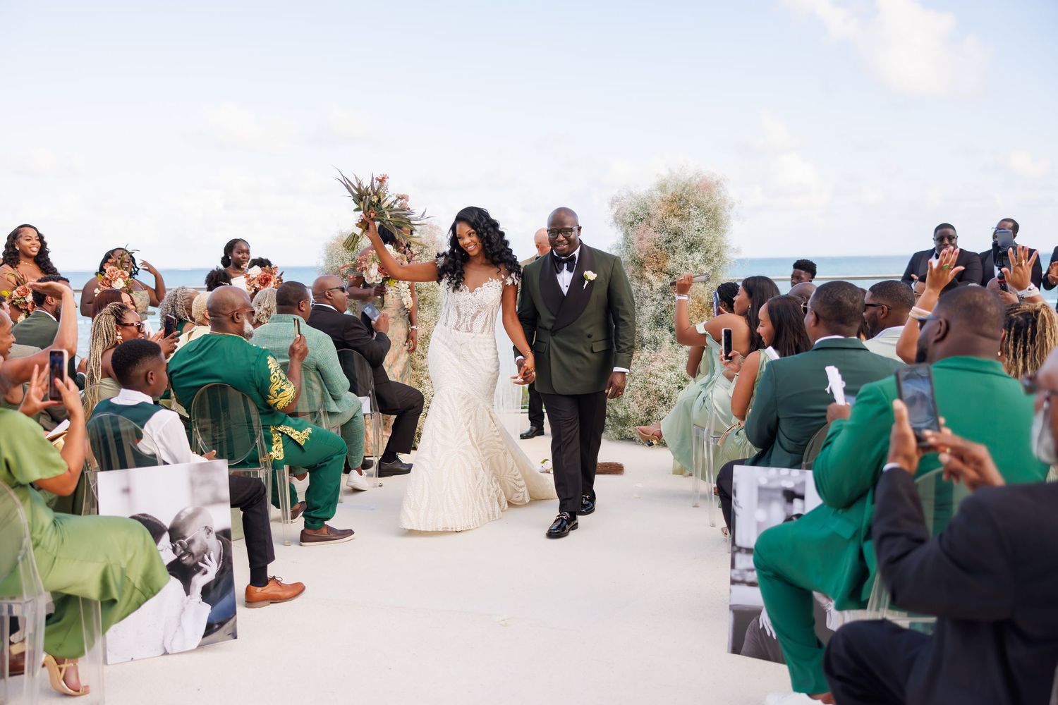 Bride and groom walk down the aisle at an outdoor wedding ceremony with guests in green attire cheering.