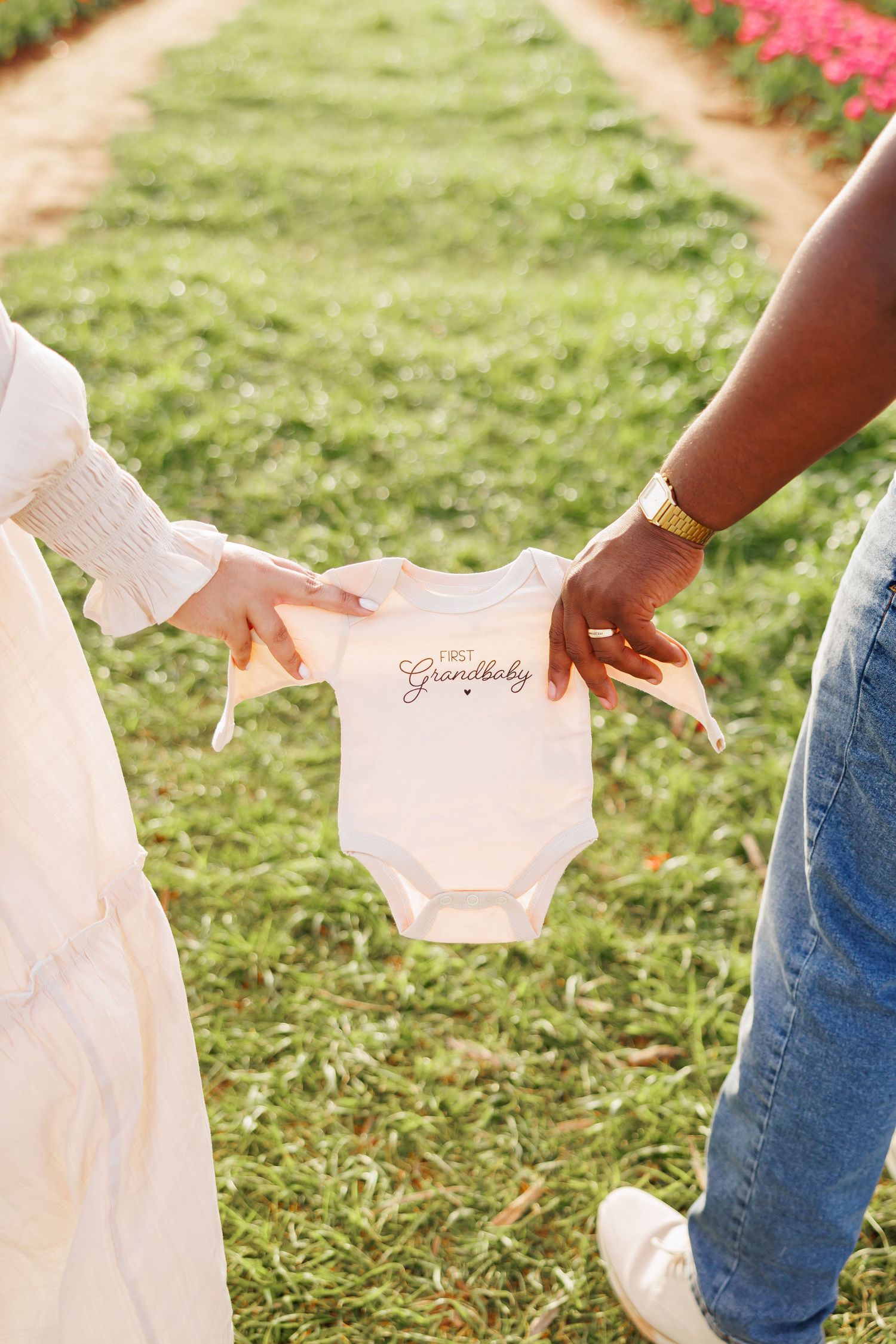 Two people holding a white 'First Grandbaby' onesie outdoors in a flower garden path.