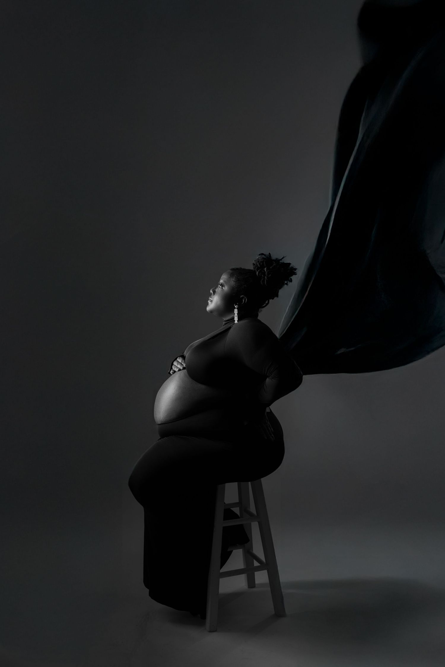 Luxury picture of A dramatic black and white maternity portrait of a pregnant woman seated on a stool with flowing fabric.