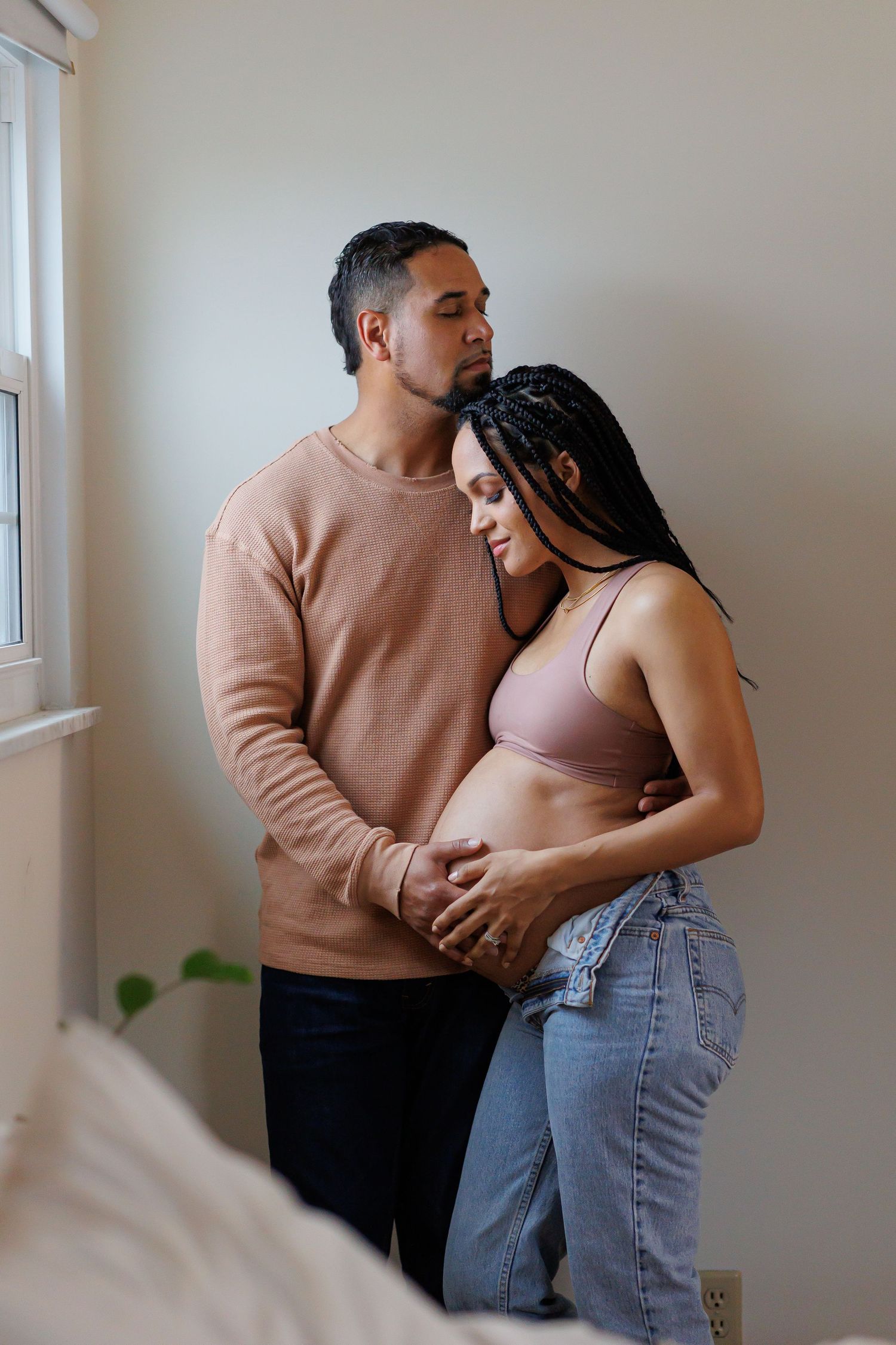 Luxury maternity picture of A couple stands by a window, man in pink sweater embracing pregnant woman in jeans and nude bralette.