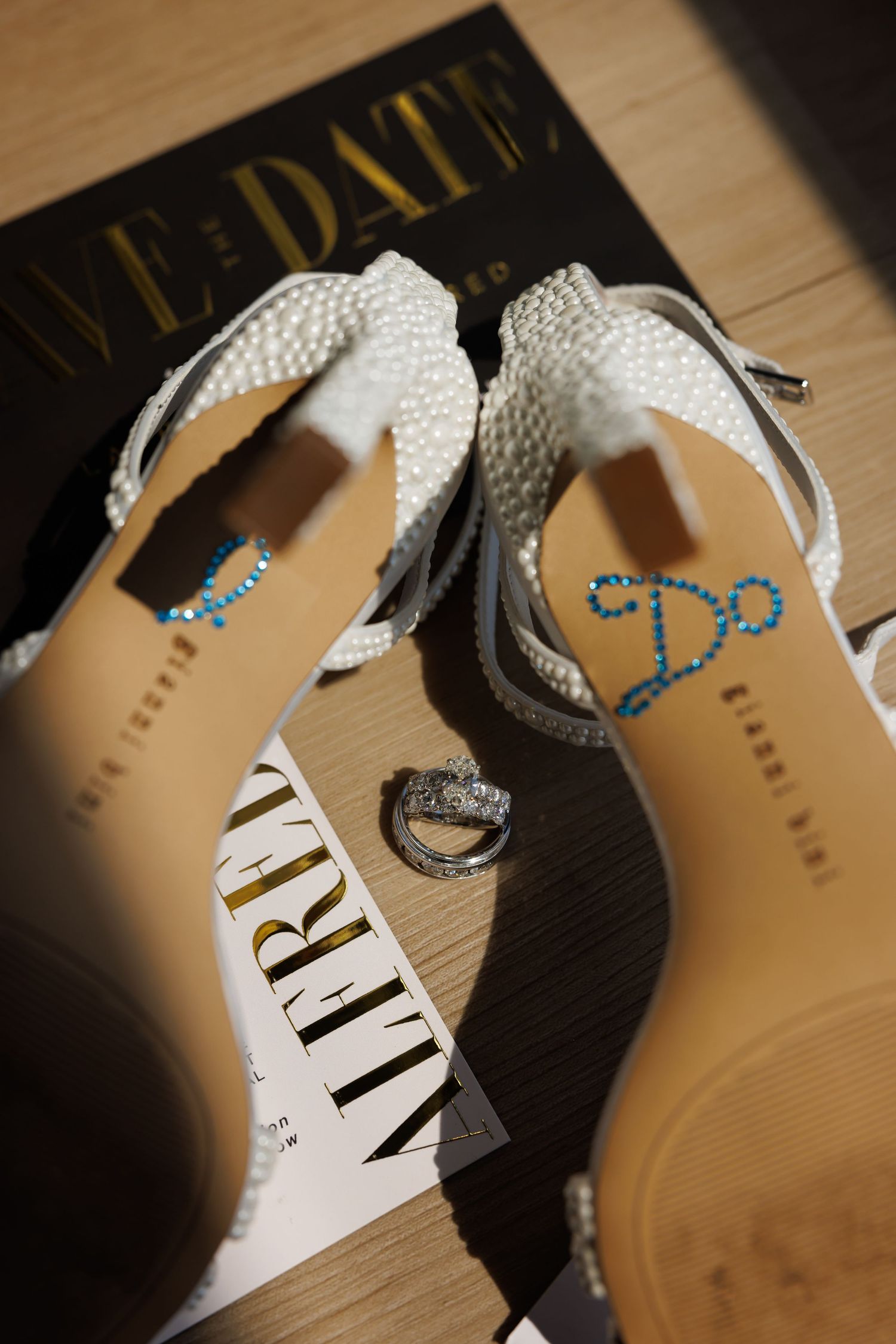 Luxury wedding picture of White pearl wedding heels with 'I Do' rhinestone insoles displayed with diamond rings on save-the-date card.