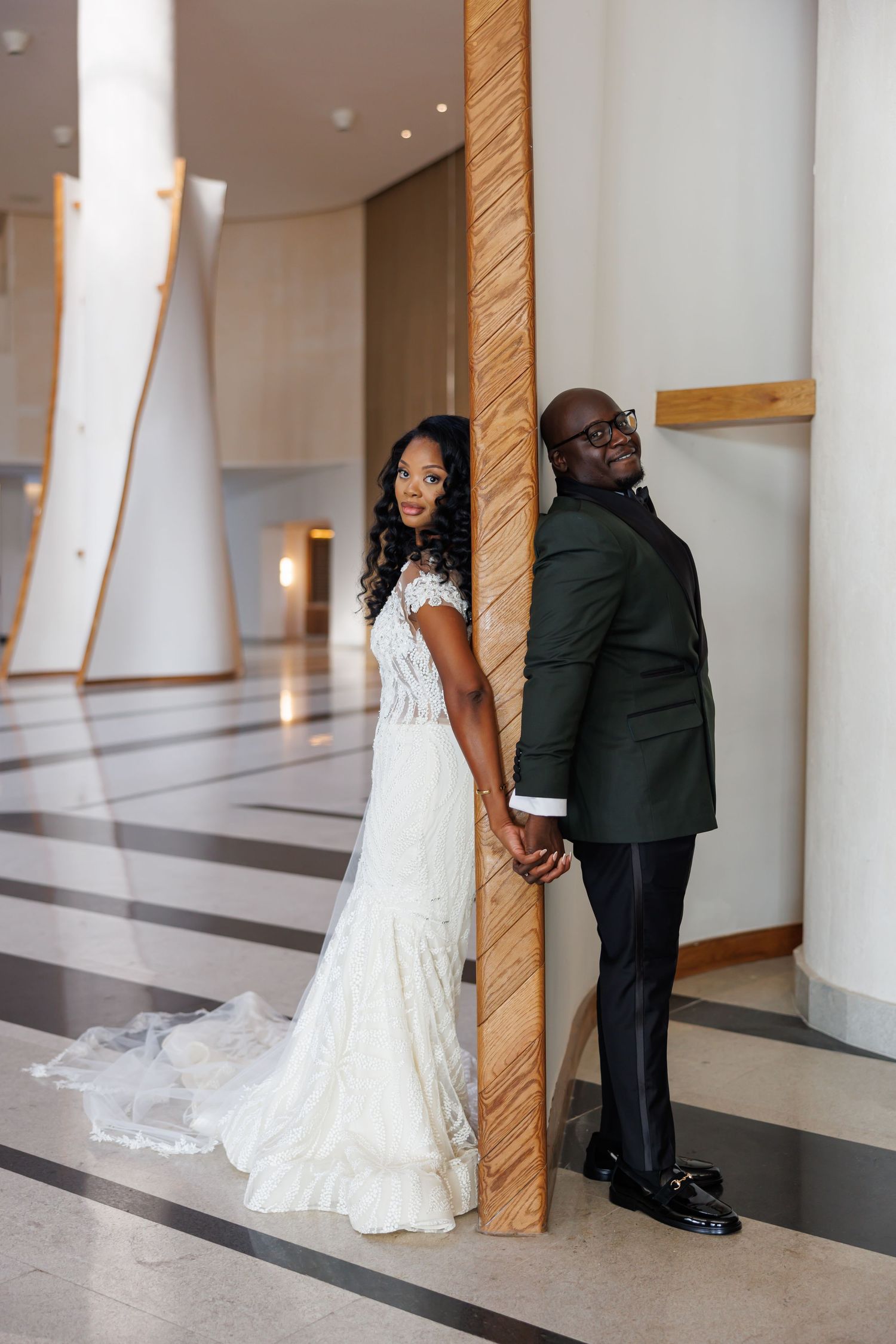 Elegant picture of bride and groom in green suit hold hands back-to-back in modern wedding venue.