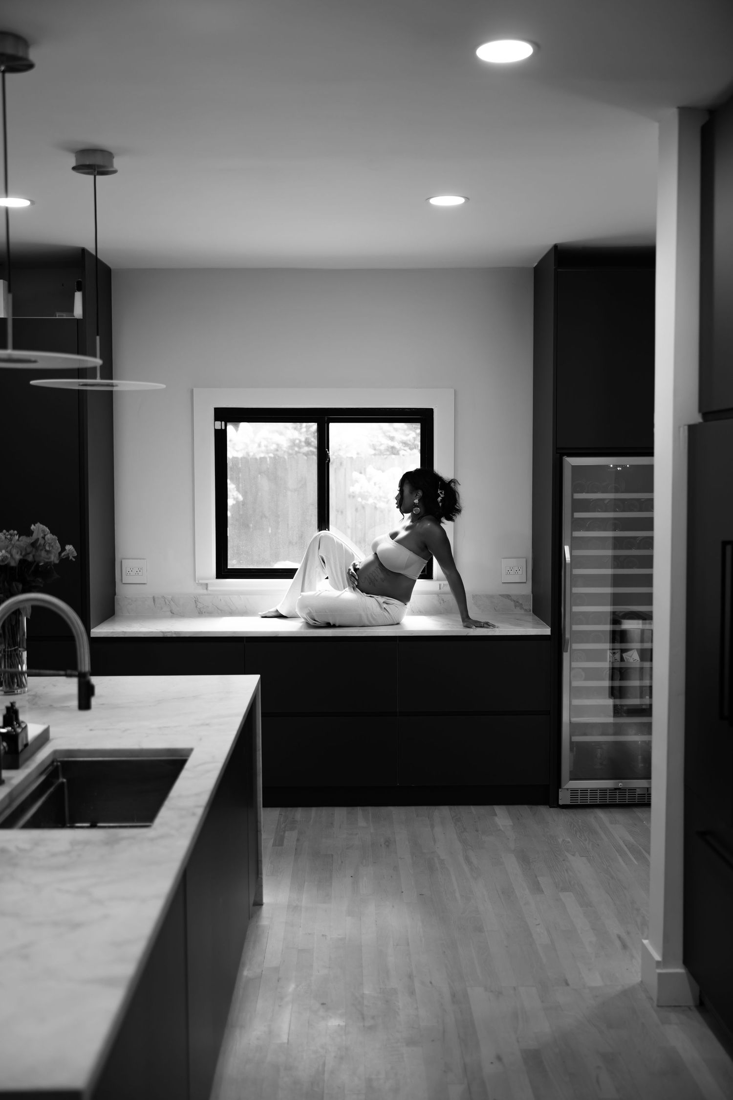 Editorial portrait of A black and white photo of a woman sitting on a kitchen counter by a window in a modern home.