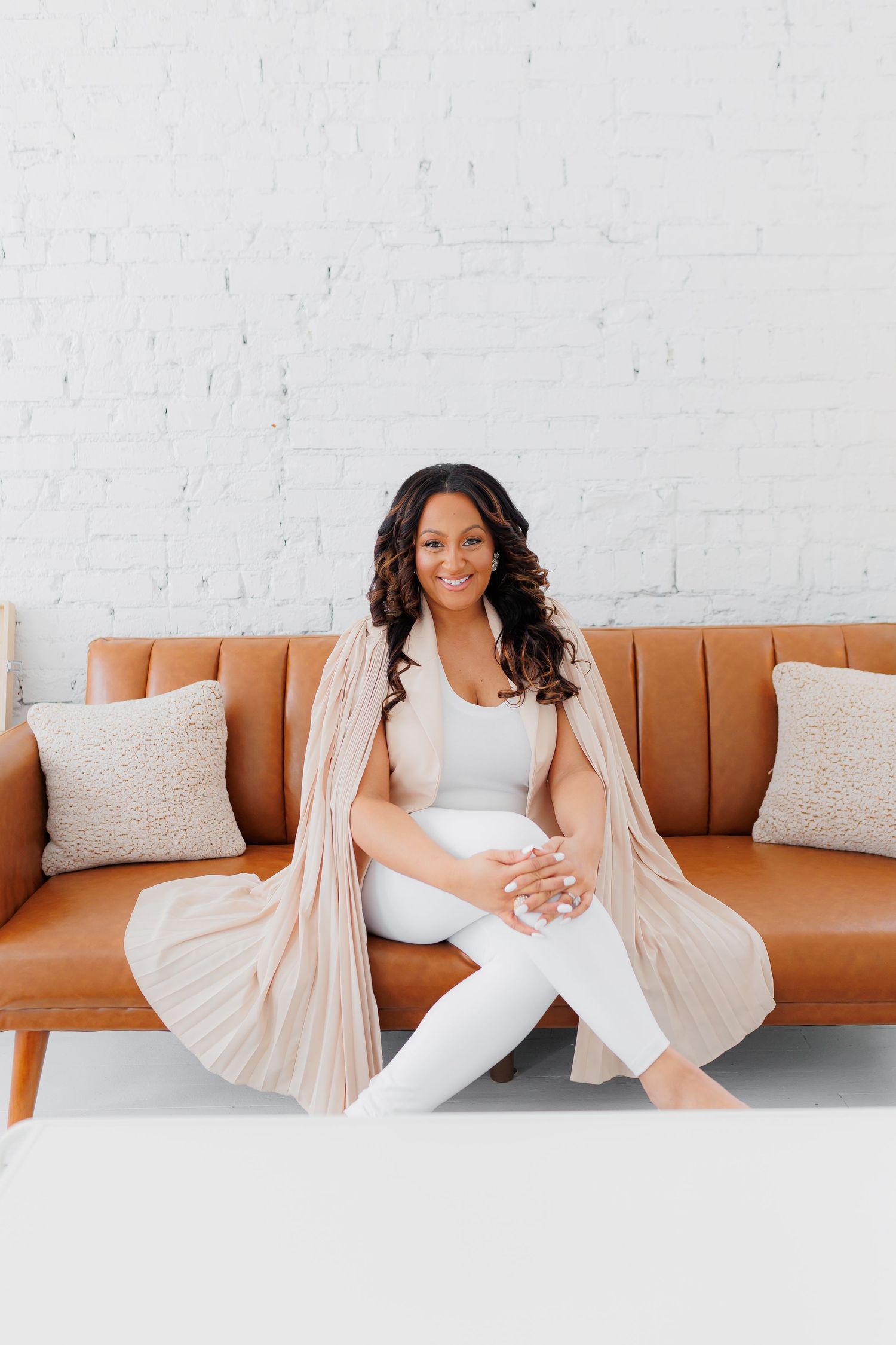 Smiling woman in white outfit and beige cardigan sitting on a tan leather sofa against a white brick wall.