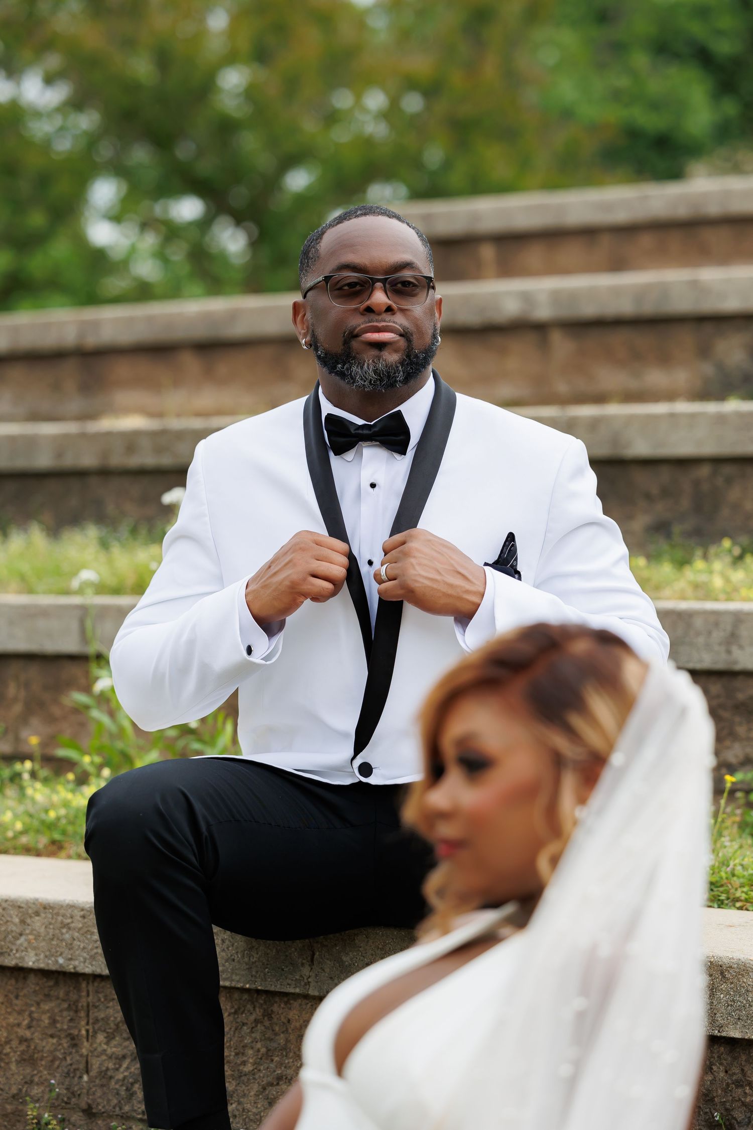 Luxury wedding picture of confident Black groom in white tuxedo poses on stone steps with bride in foreground.