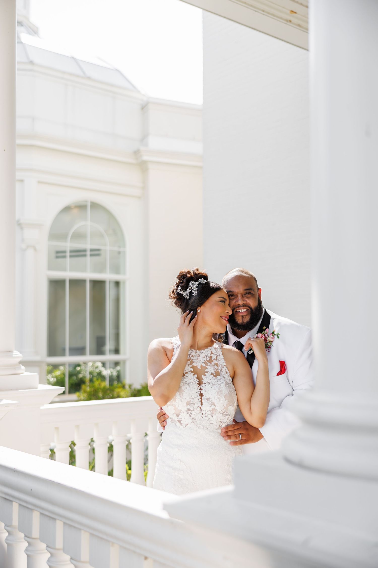 Luxury wedding picture of Smiling groom embraces bride in lace wedding dress on white columned venue balcony.
