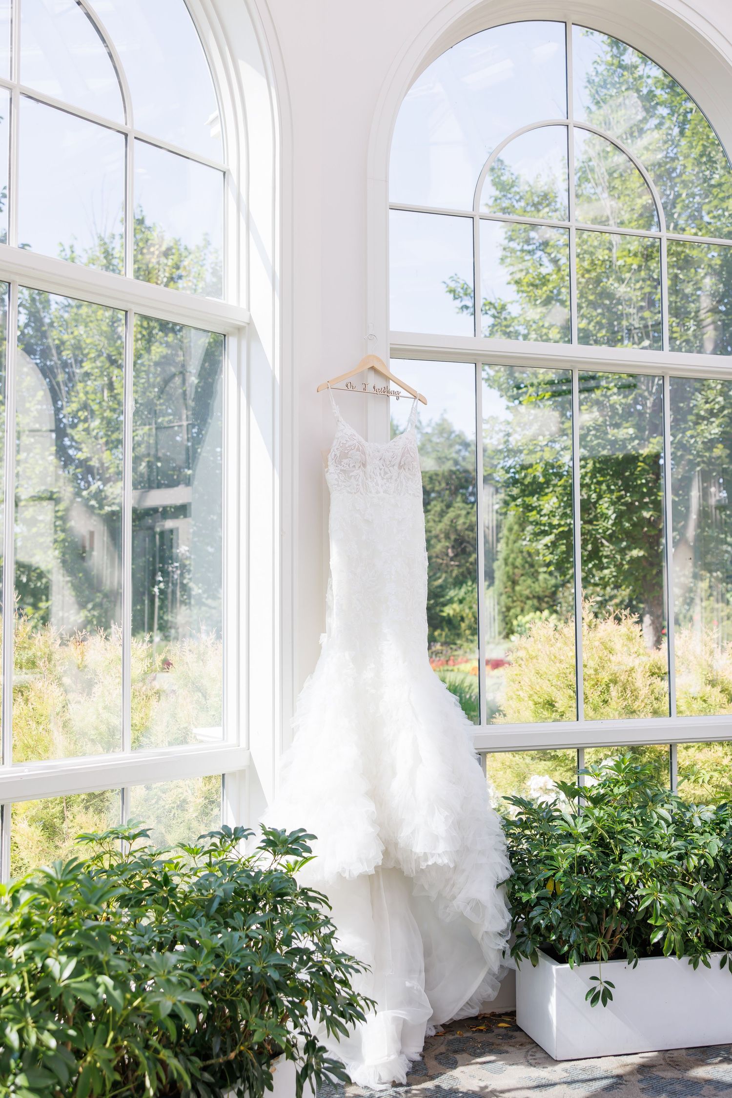 Elegant picture of White lace mermaid wedding dress hanging on a wooden hanger in a sunlit greenhouse window.