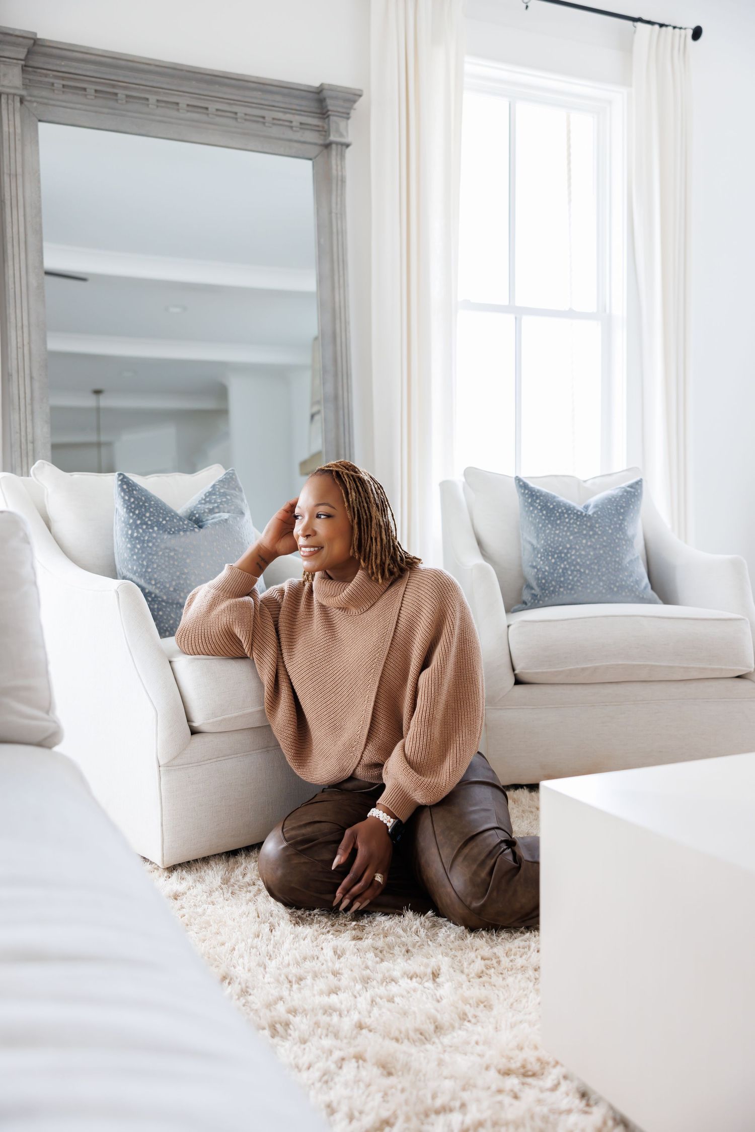 high end picture of woman in beige turtleneck sweater sitting on rug in stylish home in bright white living room.