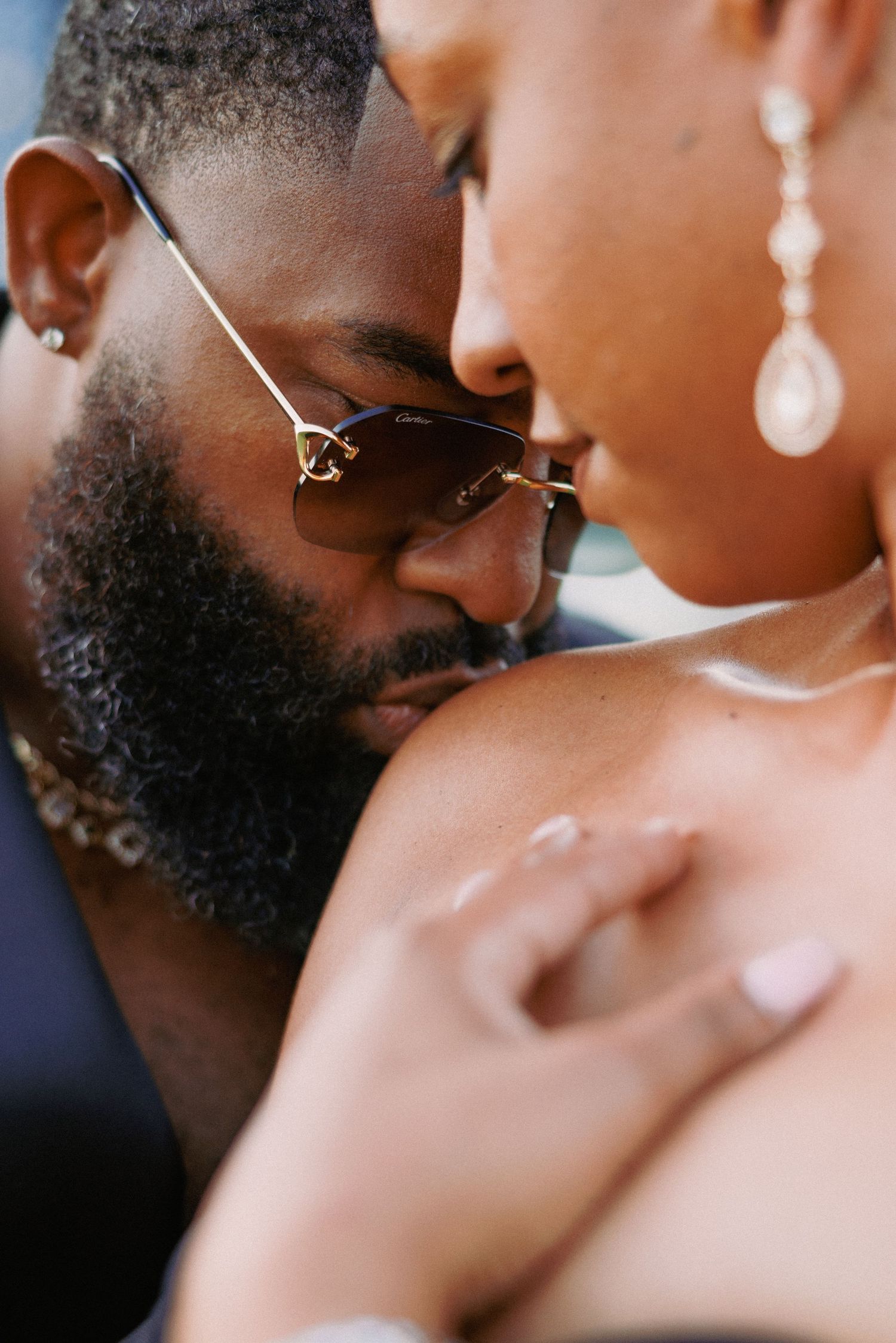A man with sunglasses and a beard gently kisses a woman's shoulder in a romantic and intimate close-up moment.