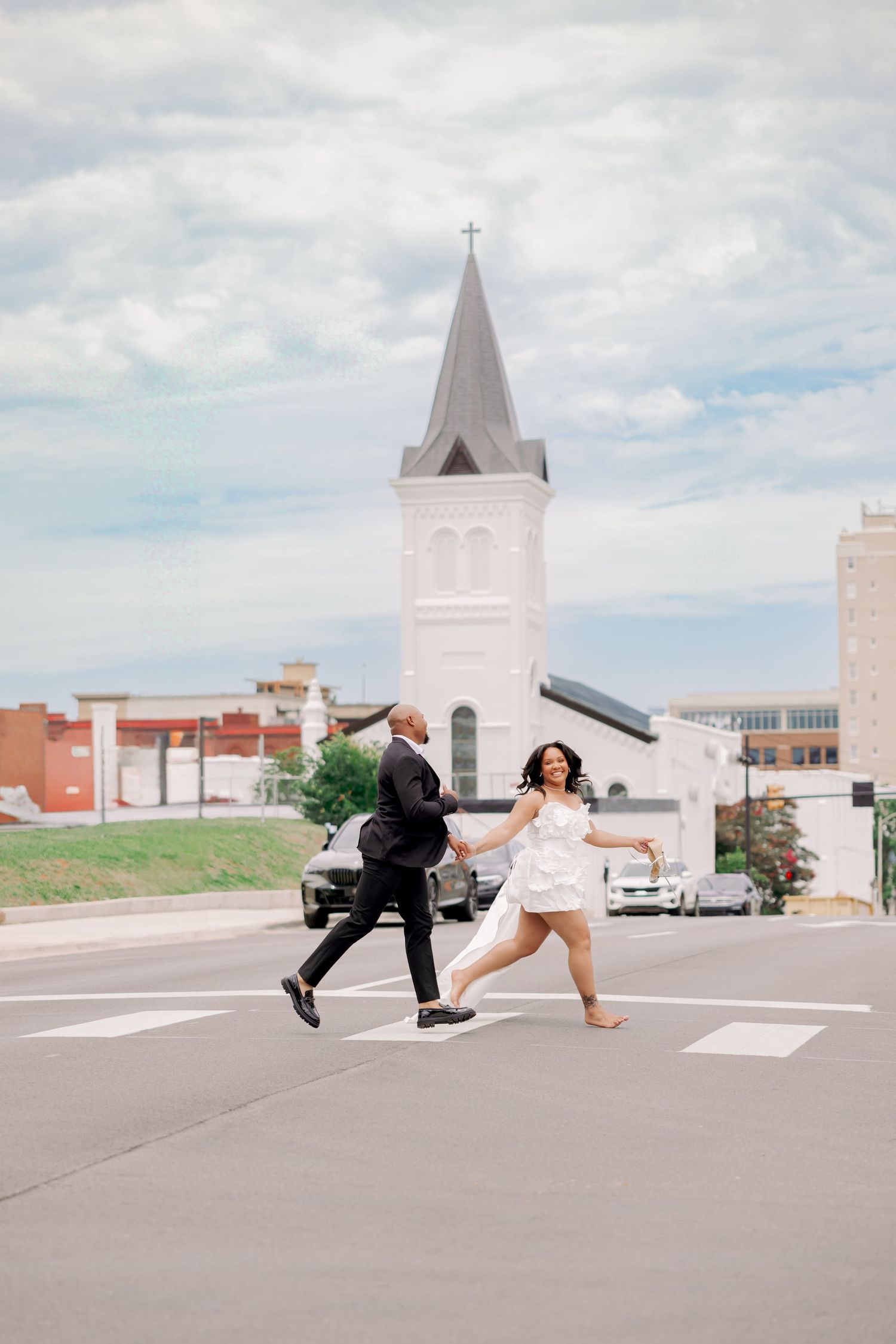 Joyful couple in wedding attire running hand-in-hand across a crosswalk with a white church steeple in the background.