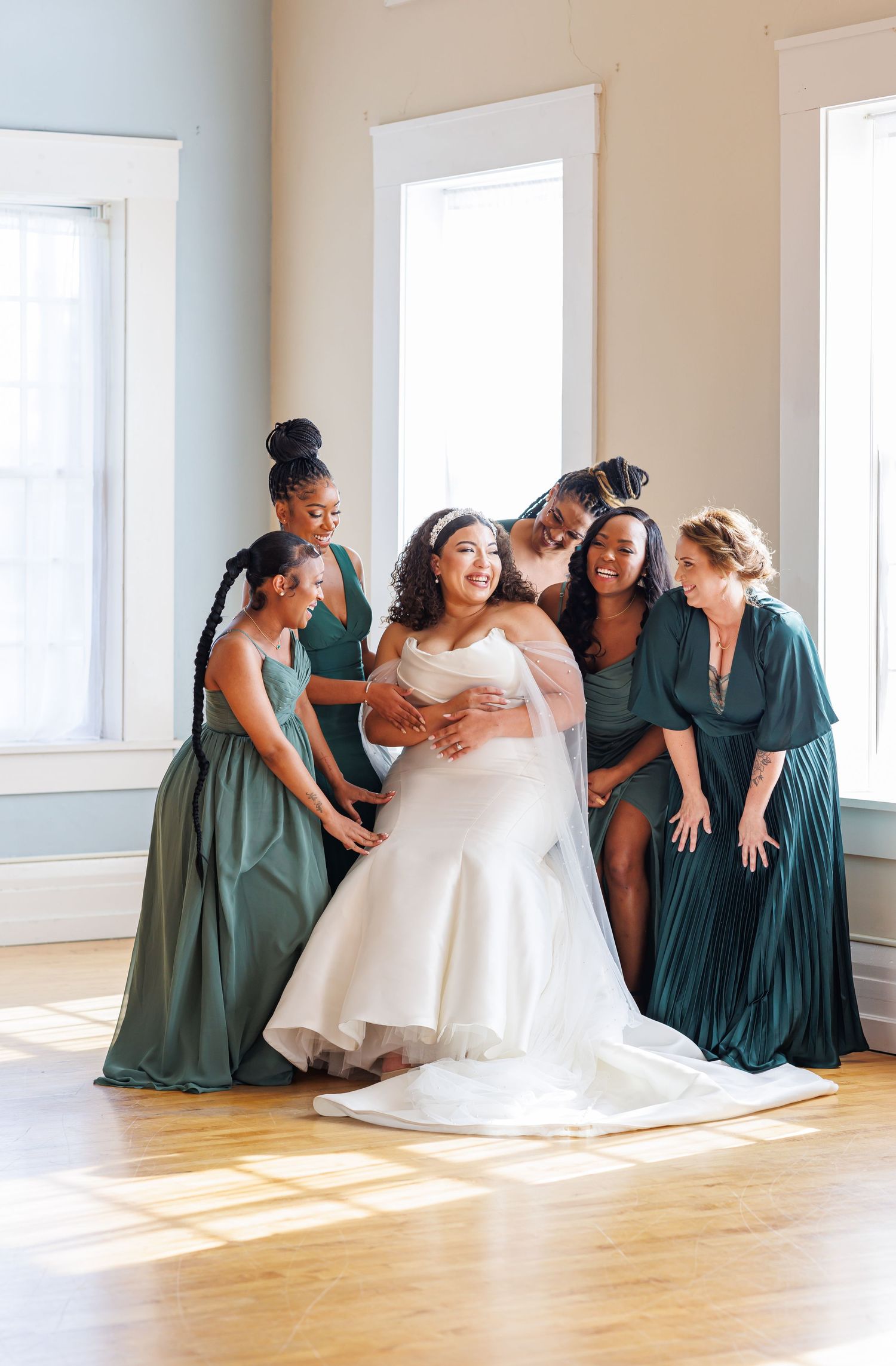 Luxury wedding portrait of Bride surrounded by bridesmaids in emerald green gowns sharing a joyful moment in a sunlit room.