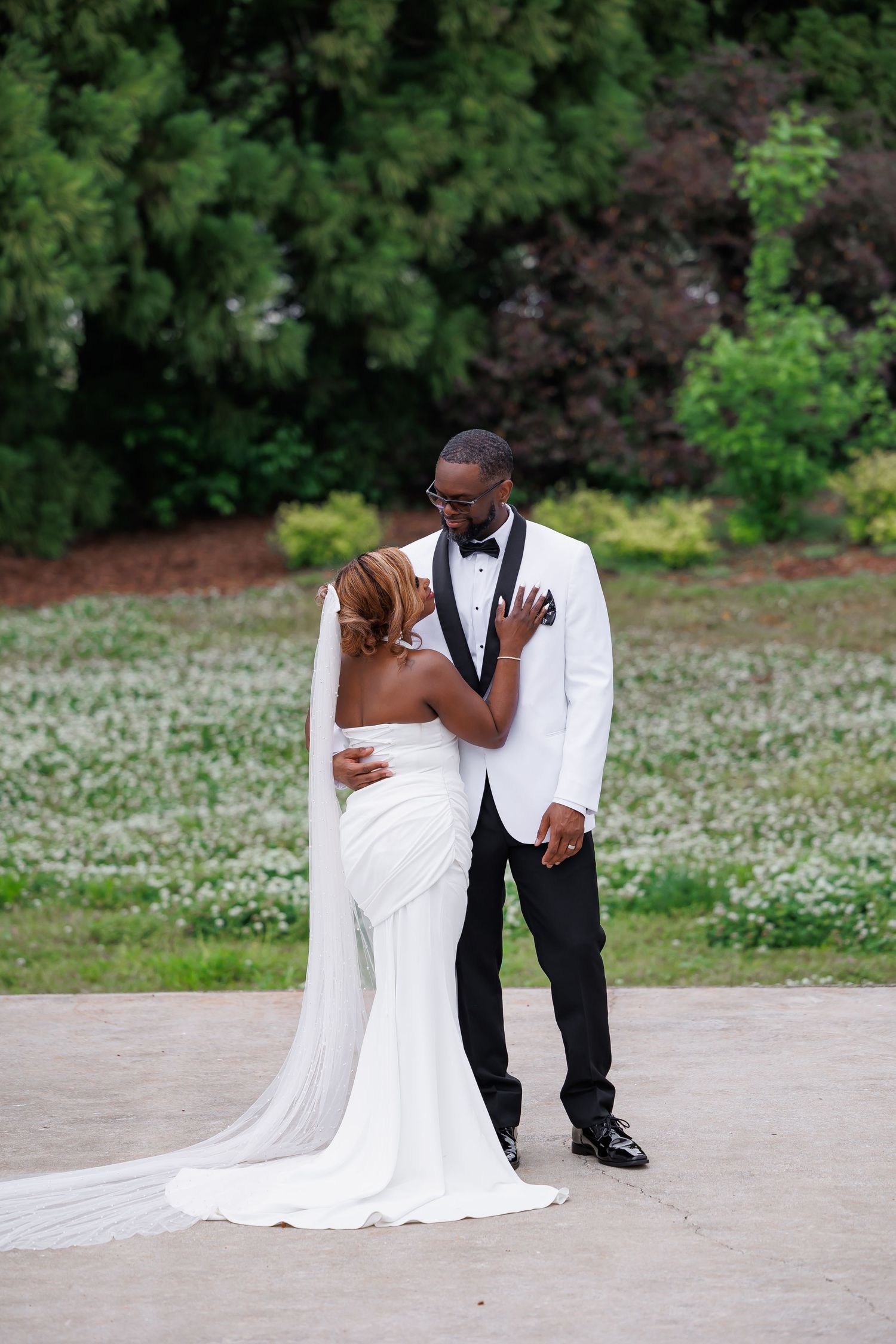 Luxury wedding picture of Bride and groom share an intimate moment in white wedding attire on a floral garden patio.