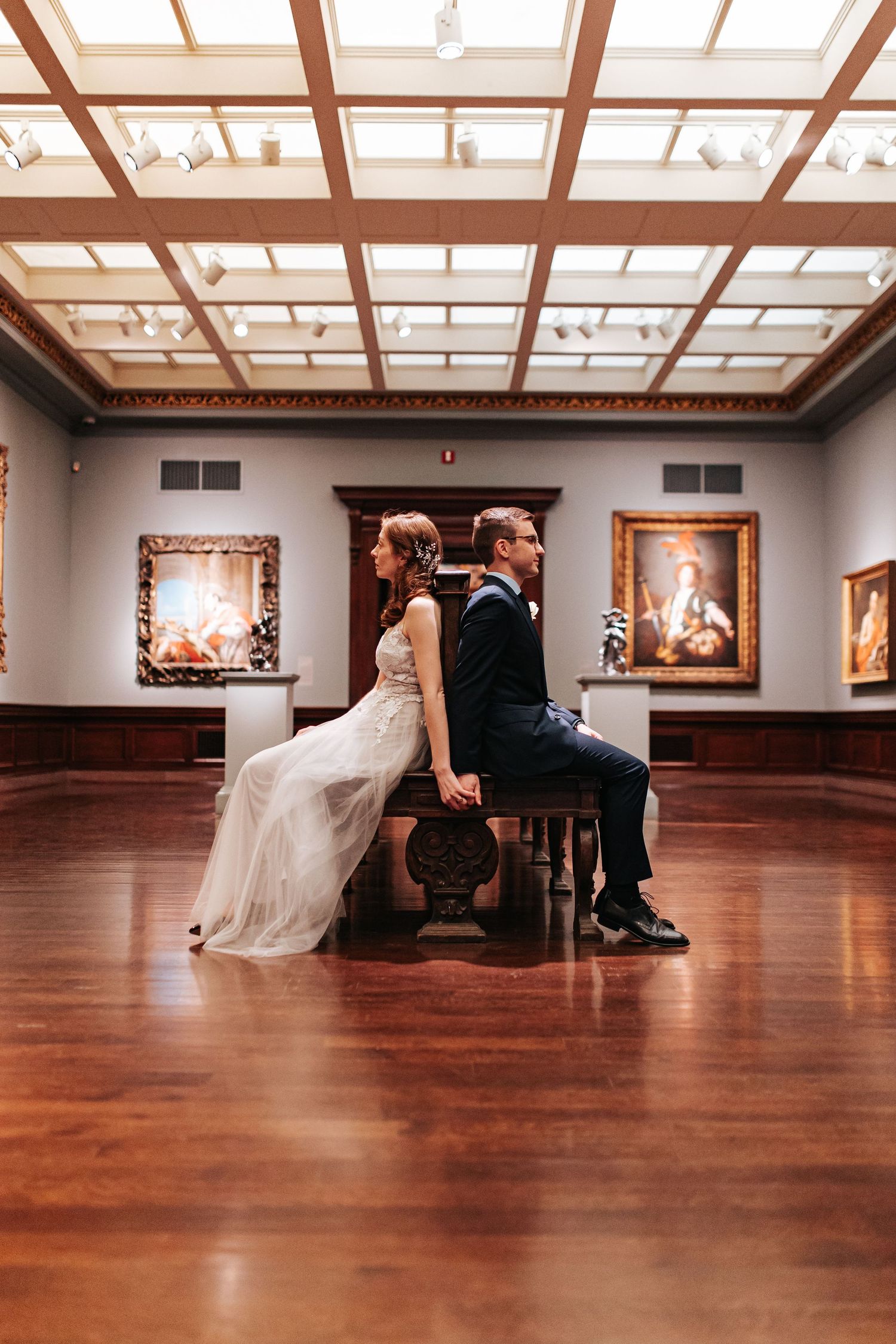 Bride and groom sitting back-to-back on a bench in an elegant art museum gallery with hardwood floors.