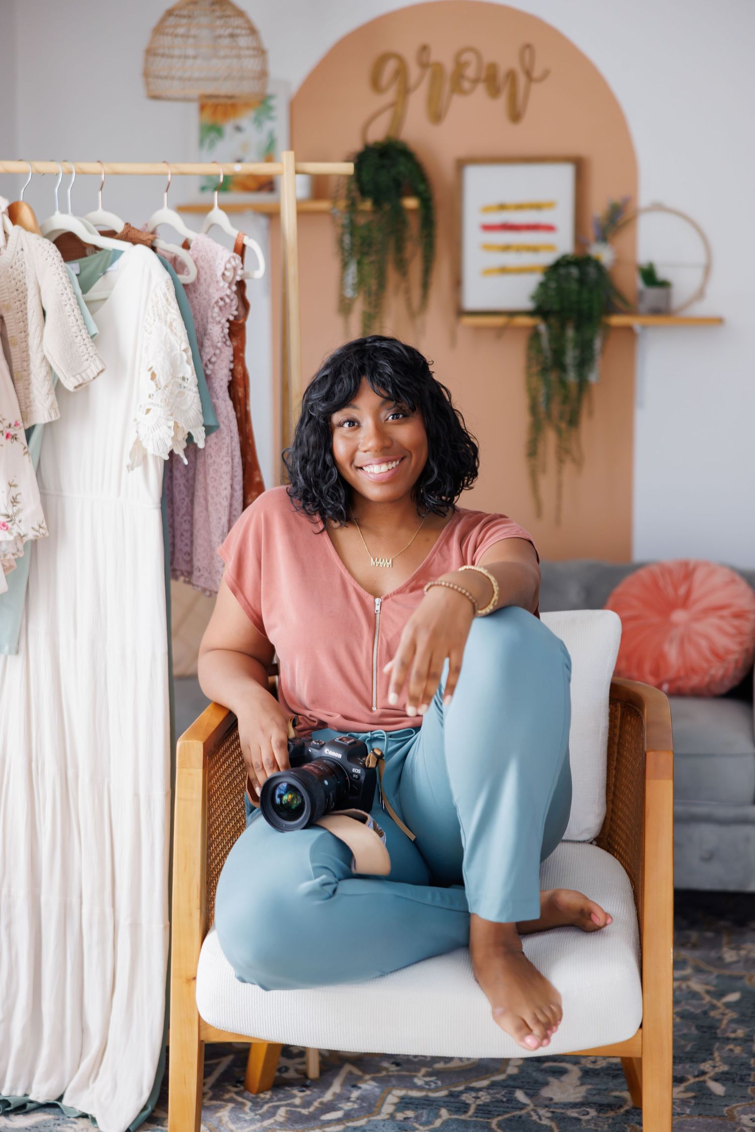 Natural picture of Smiling Black female photographer sitting cross-legged in chair holding camera in boho studio.