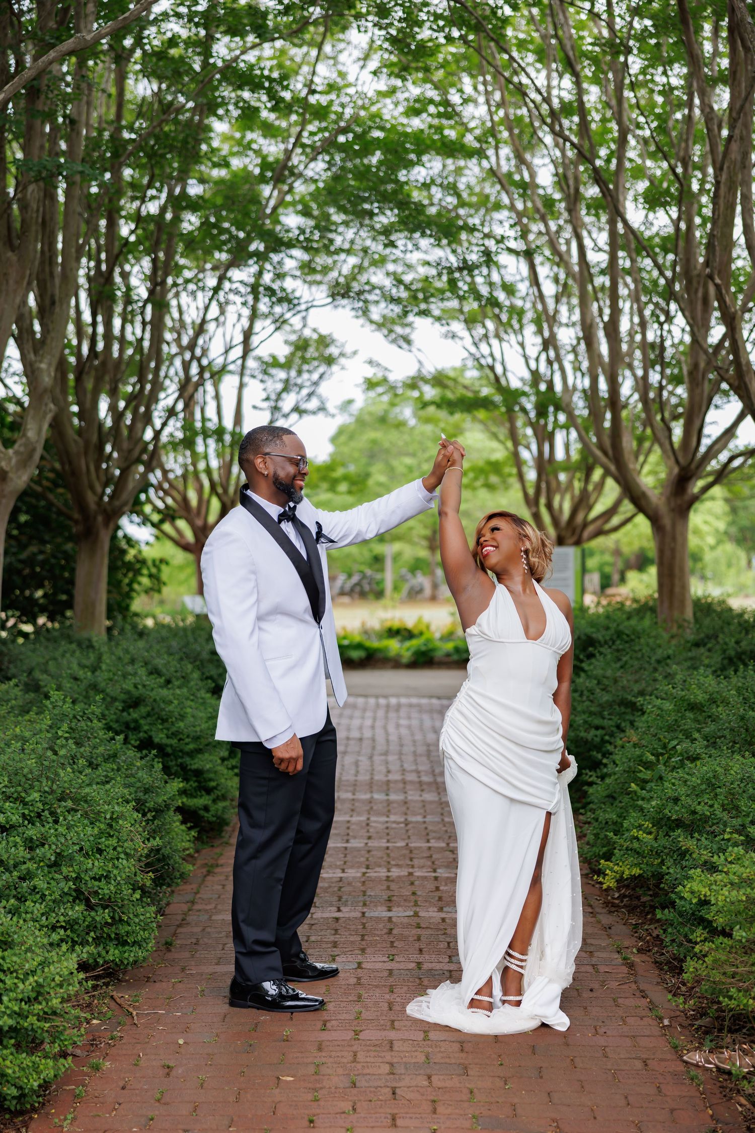 Luxury wedding picture of groom twirls bride in white wedding attire along a brick pathway lined with lush green trees.