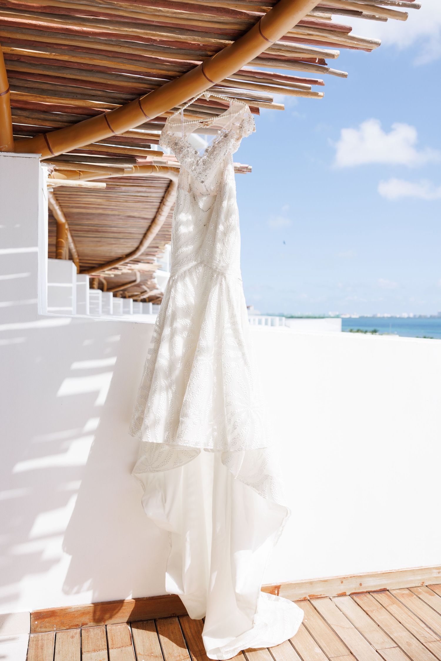 White lace wedding dress hanging on a wooden pergola with ocean view on a sunny day.