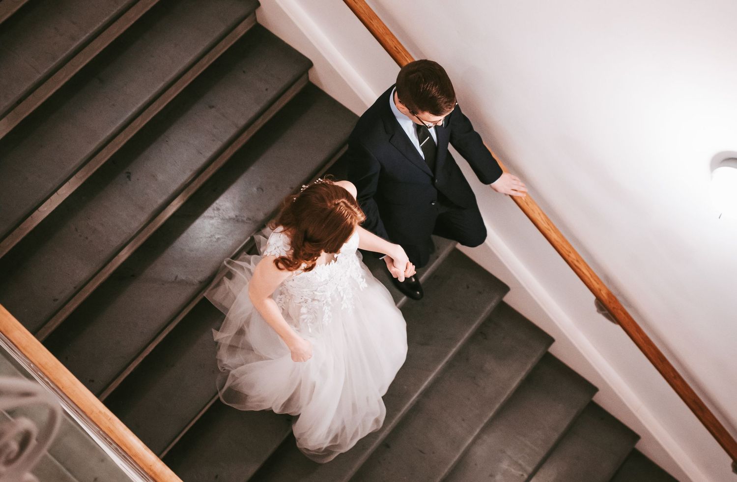 Bride and groom walking down elegant staircase, aerial view of wedding couple in white gown and black suit.