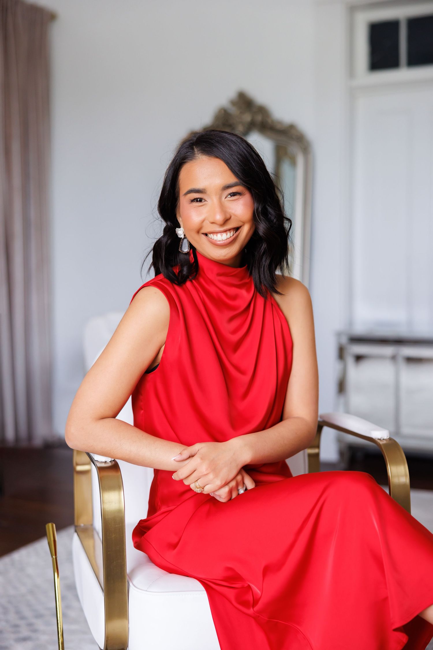 High-end modern picture of Smiling woman in red sleeveless dress seated in gold accent chair in elegant room.