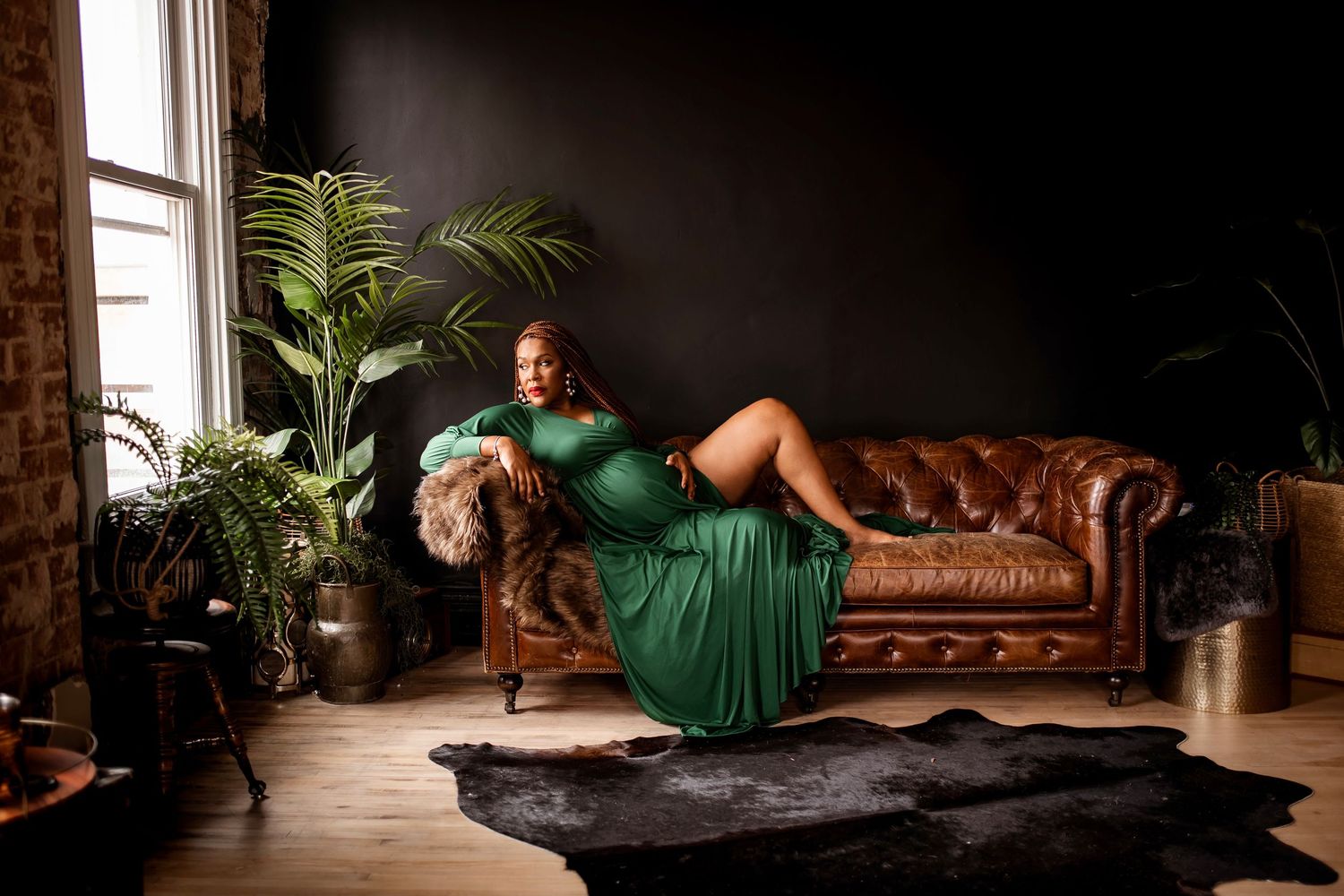 Plus-size woman in elegant green gown reclines on brown leather chesterfield sofa in dark, moody studio setting.