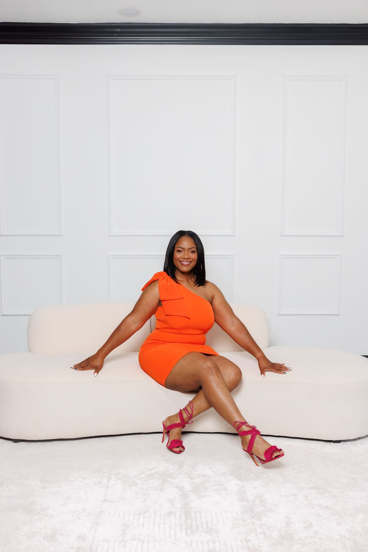 Luxury picture of Woman in orange one-shoulder mini dress and pink heels sitting on white sofa smiling.