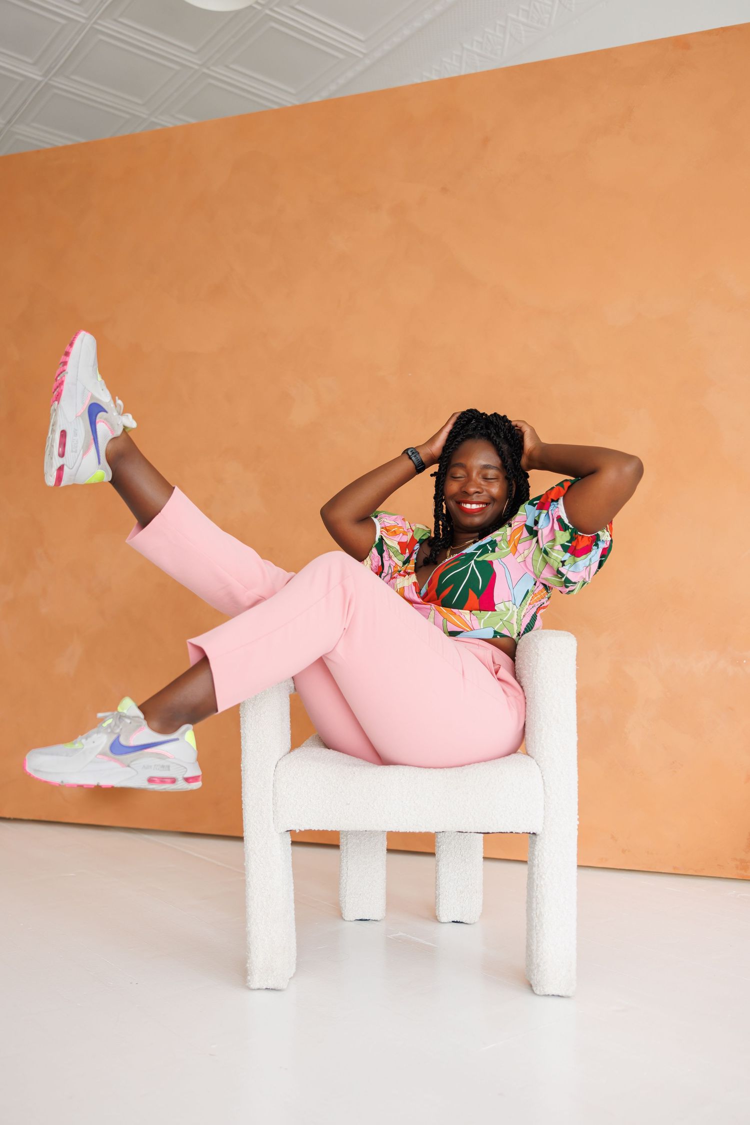 High-end picture of Woman in floral top and pink pants relaxing in white chair against orange wall.