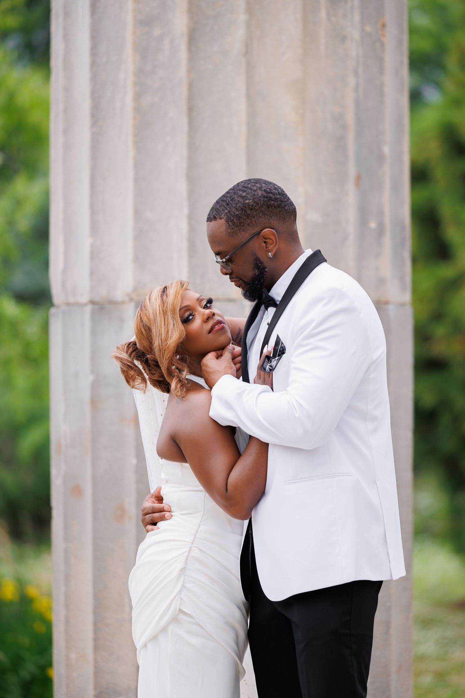 High end portrait of Black groom in white tuxedo tenderly embraces bride in sleeveless wedding gown near rustic stone columns.