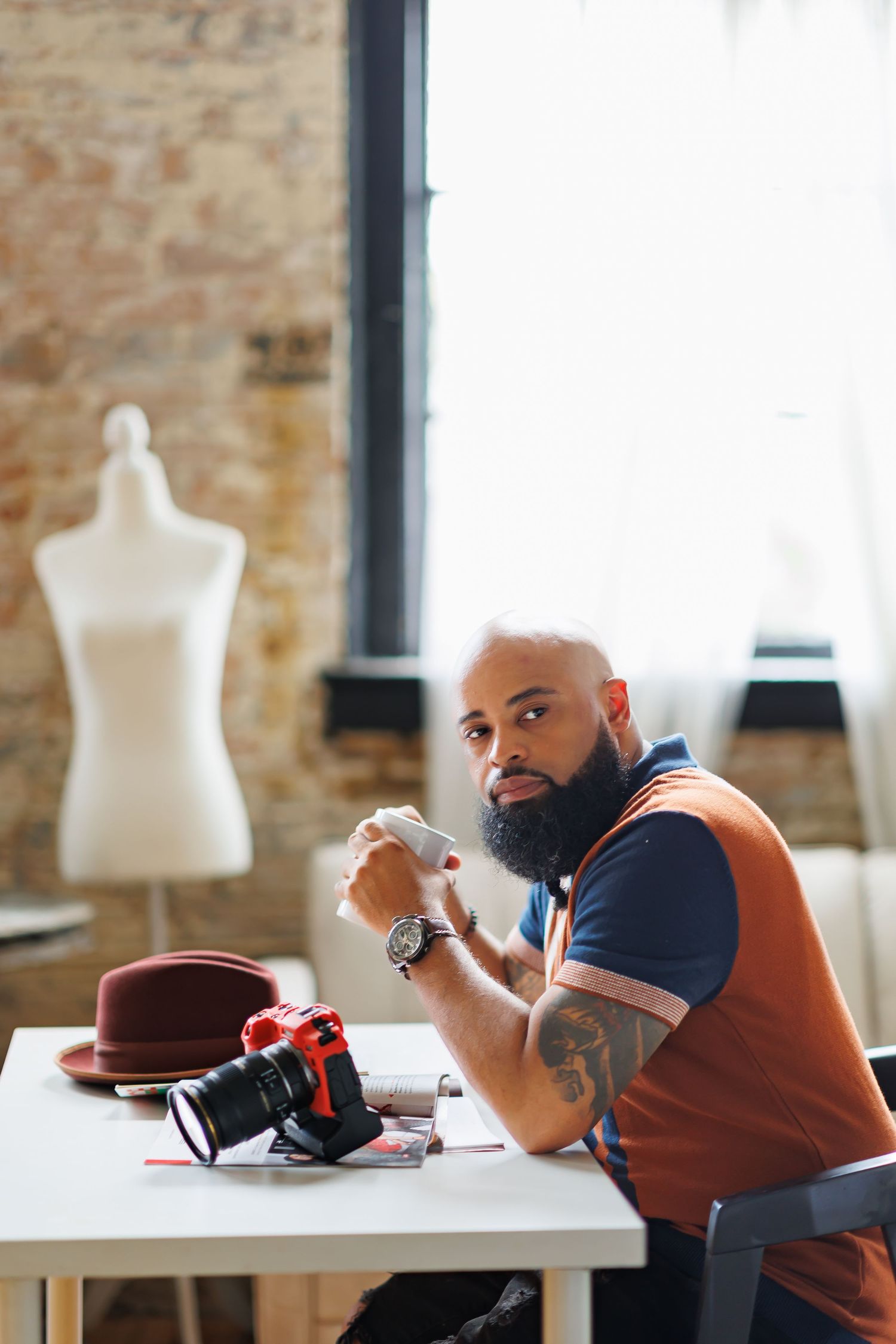 Branding picture of Bearded tattooed man sitting at desk with camera and hat in creative studio workspace.