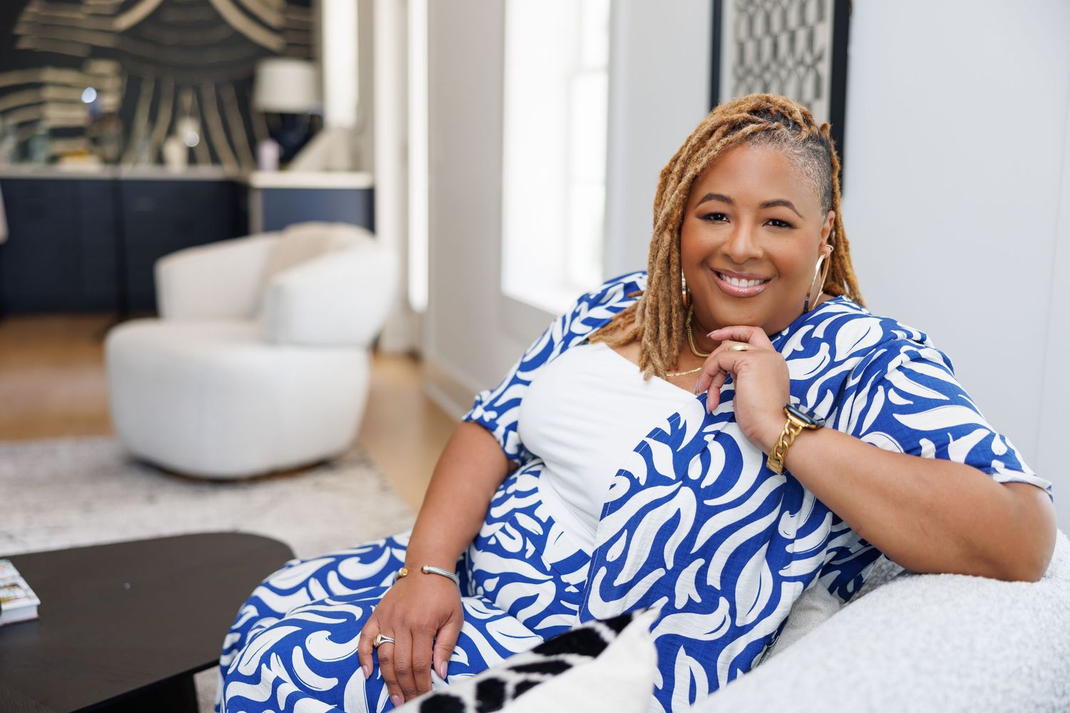 Cheerful plus-size Black woman in blue floral dress relaxing on white sofa in a modern living room. Natural light