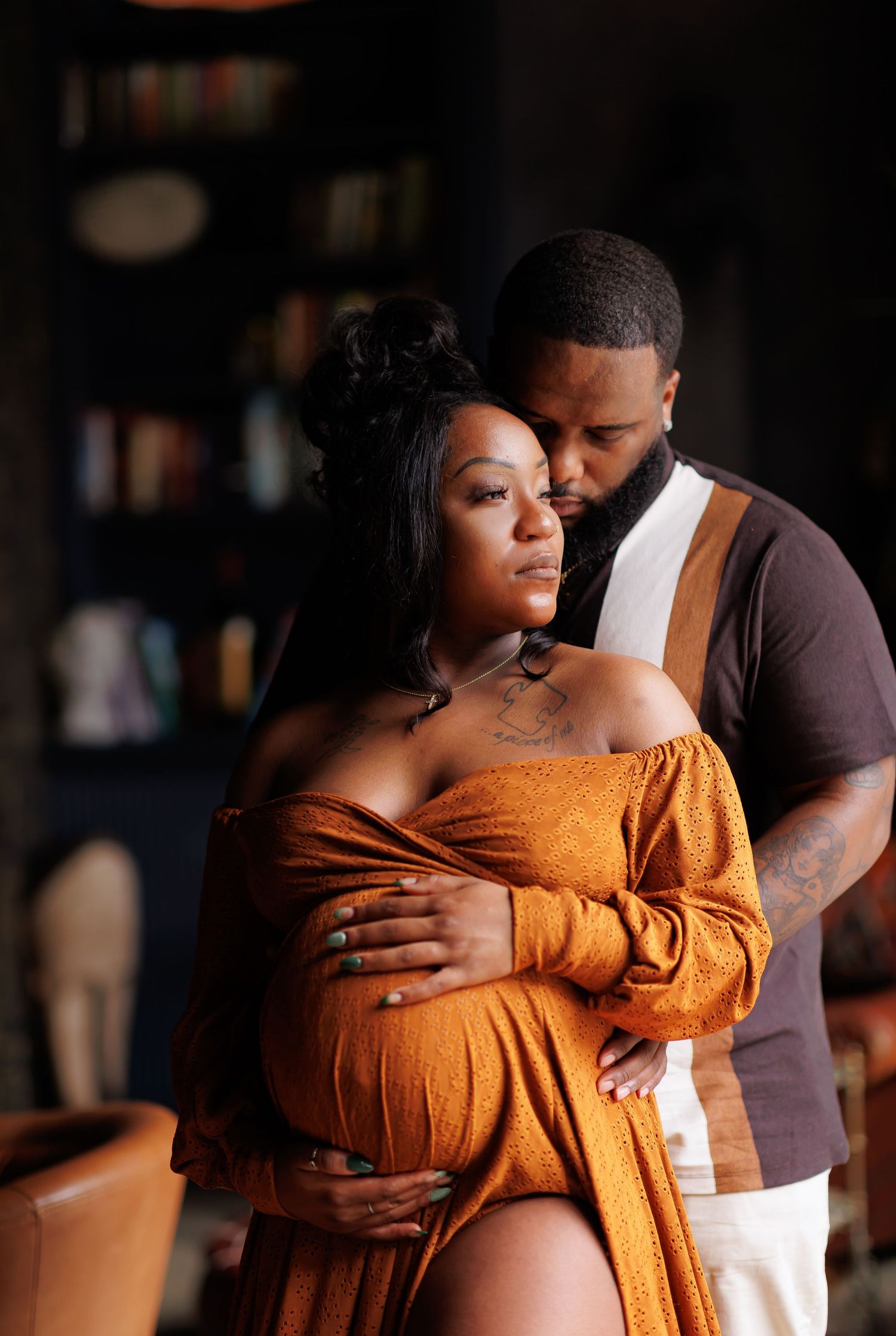 High-end portrait of A couple shares a tender maternity moment, man embracing pregnant woman in rust orange off-shoulder gown.
