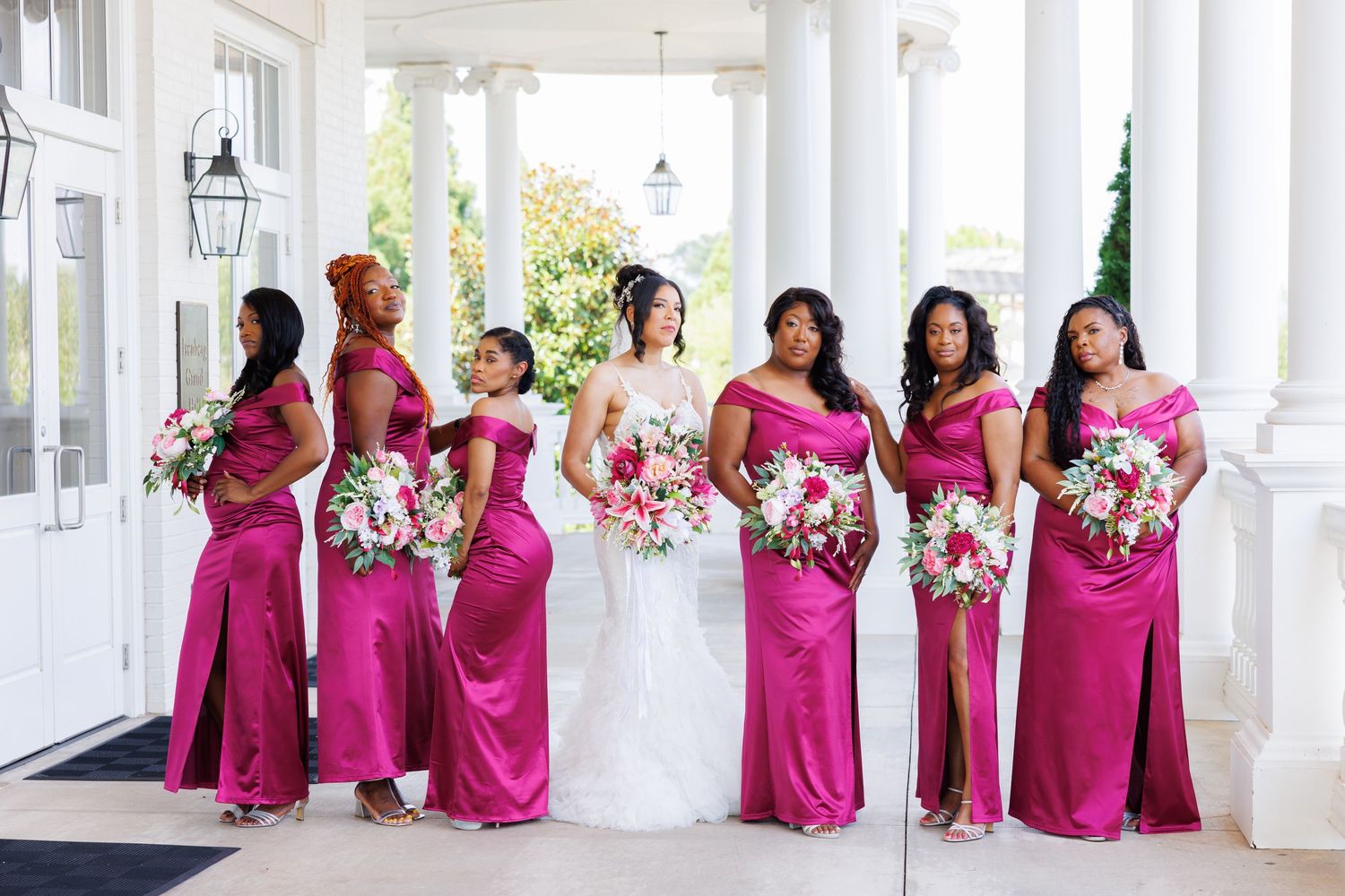 Luxury wedding picture of Bride with bridesmaids in fuchsia satin gowns holding bouquets under white columns outdoors.