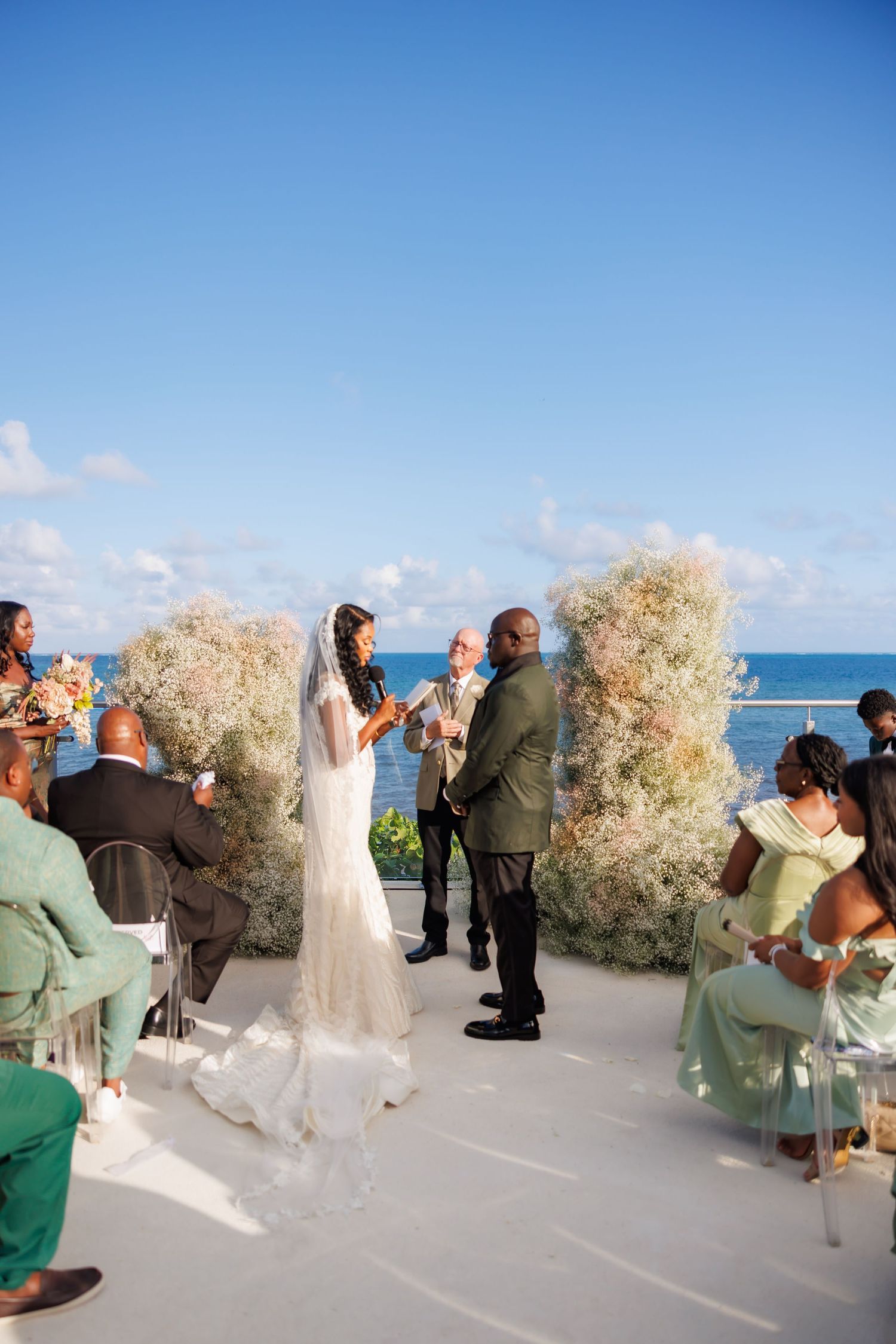Editorial picture of Bride and groom exchange vows at an outdoor oceanfront ceremony with floral arch backdrop.