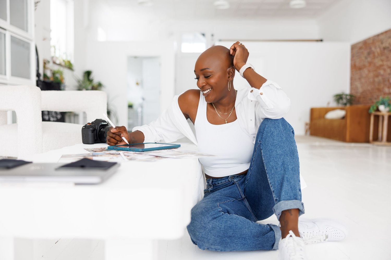 Timeless studio portrait of Bald Black woman in white top and jeans sitting on floor reviewing photos with camera nearby.