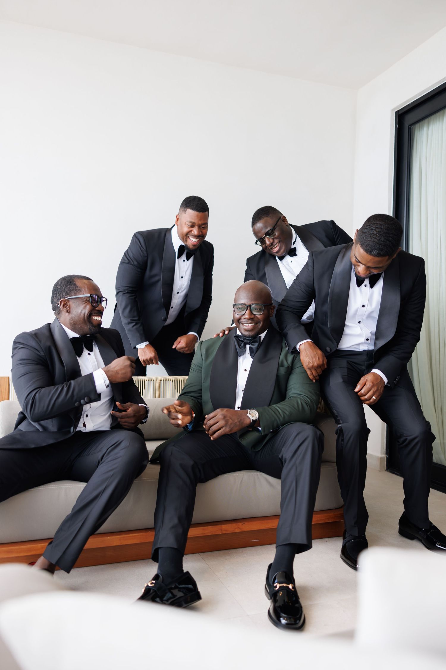 Five men in black tuxedos laughing together on a couch, with one groom in a green vest celebrating before his wedding.