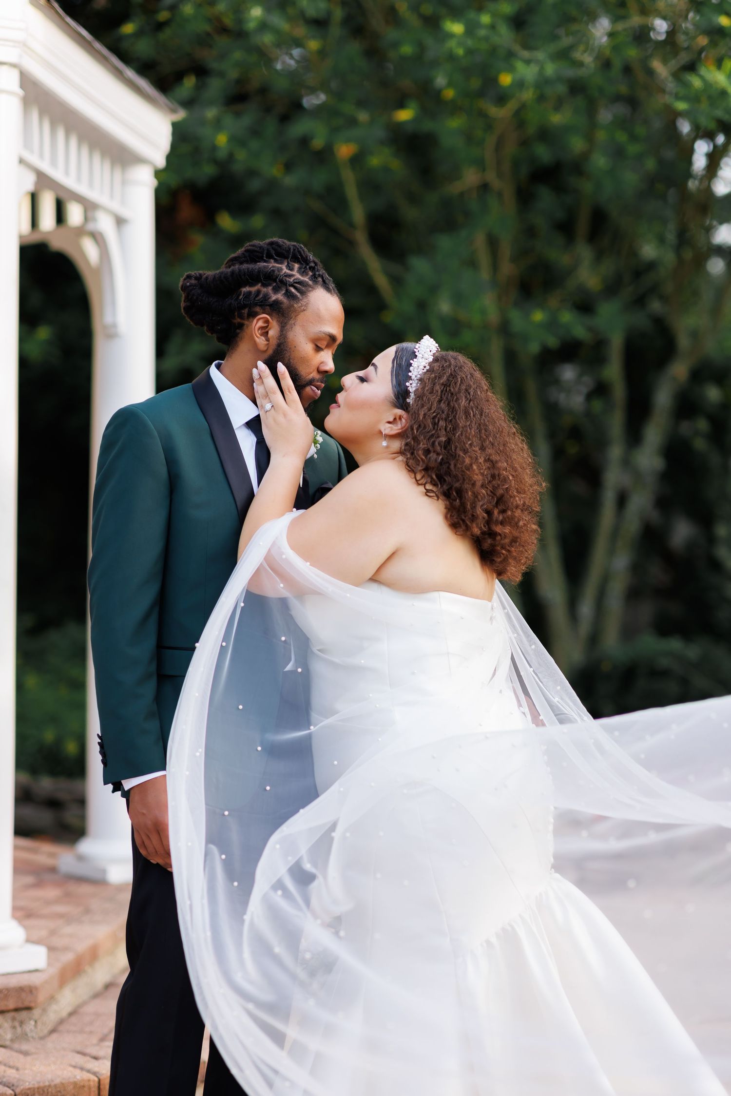 Luxury wedding picture of Bride and groom share a romantic moment near a white gazebo as her veil billows in the wind.