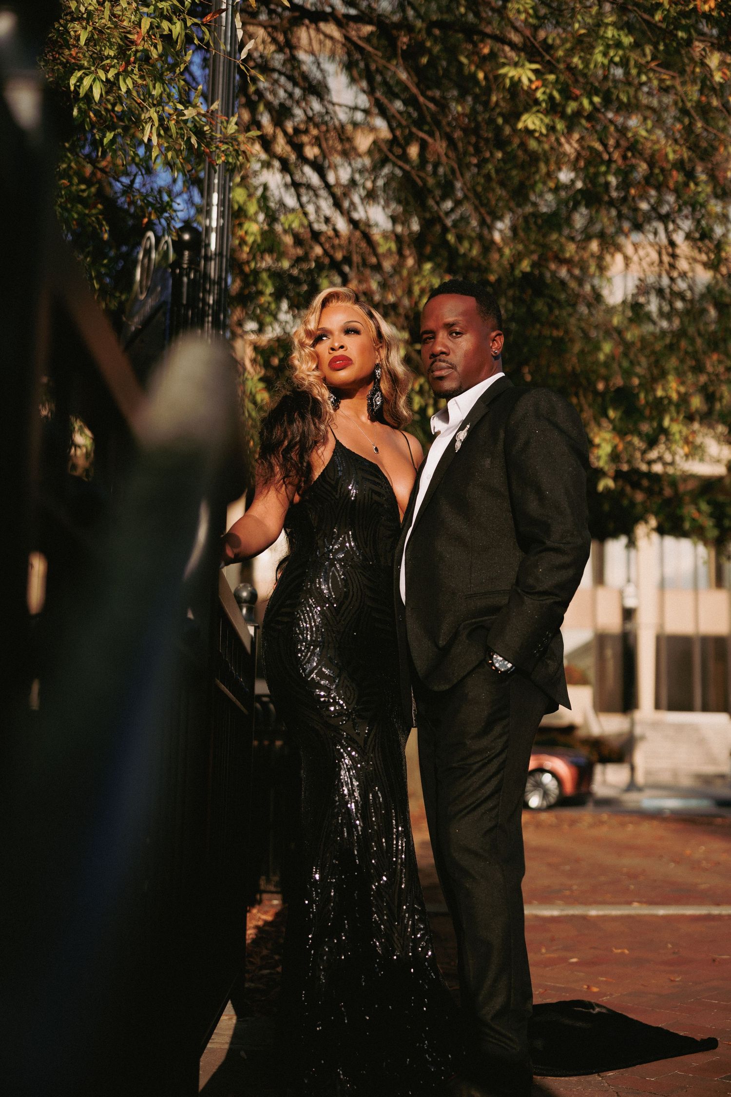 Glamorous picture couple posing outdoors at dusk, woman in black sequin gown, man in black suit.