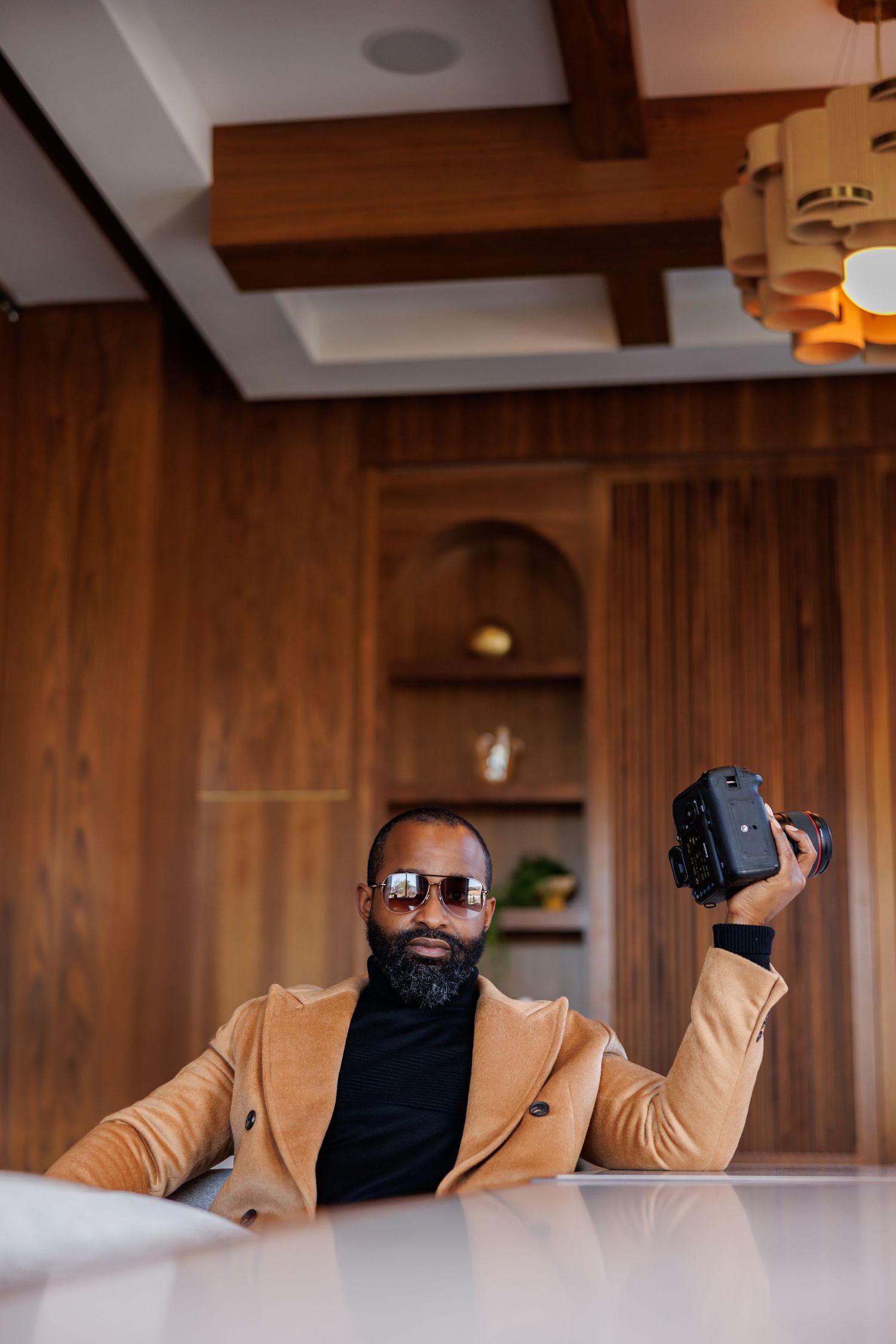High end portrait of stylish Black man in tan blazer holds camera up in a warm wood-paneled room.