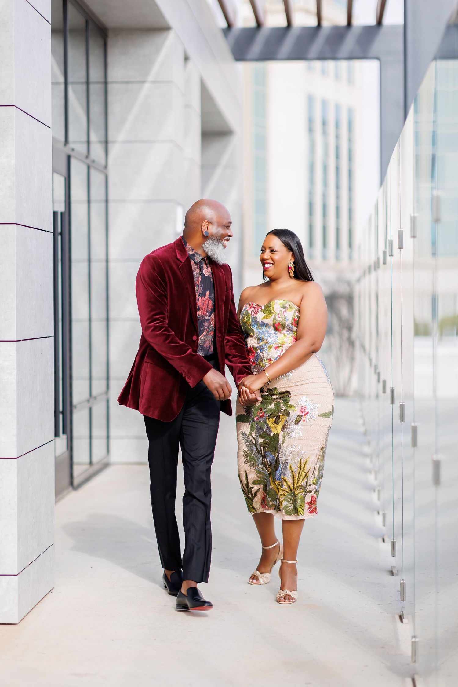 Elegant picture of Couple walking hand in hand outside modern building, man in burgundy velvet blazer, woman in floral midi dress.