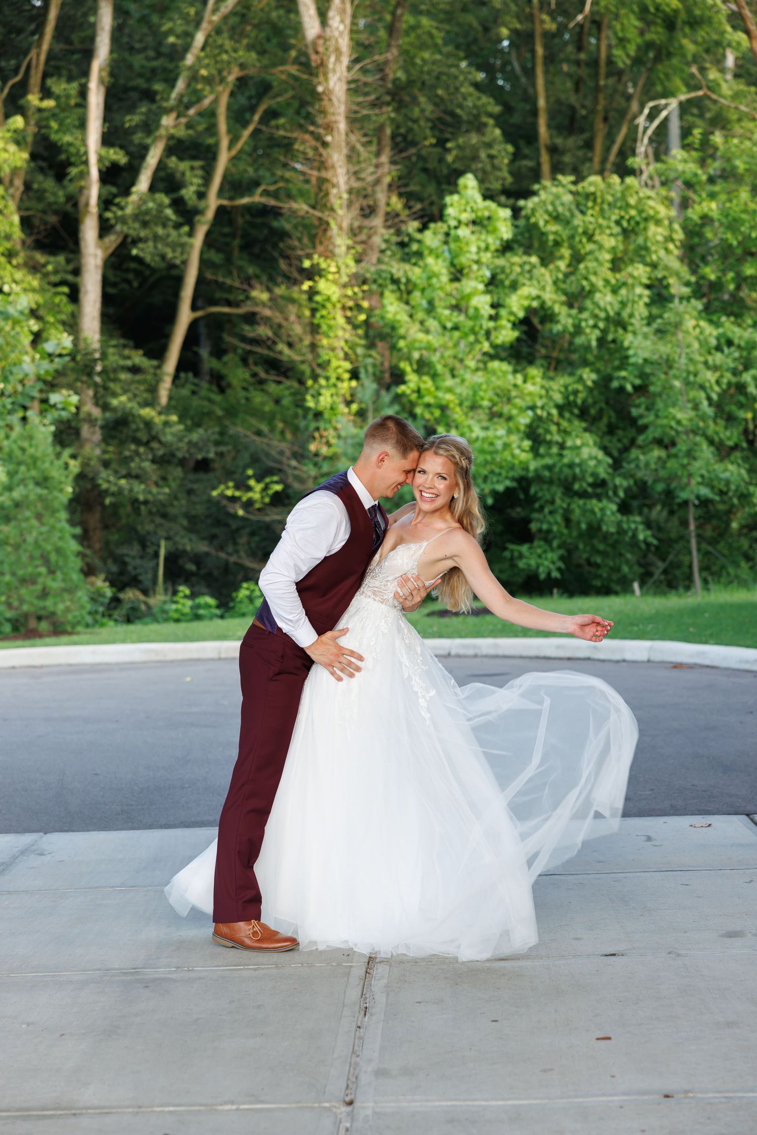 Luxury wedding picture ofBride in flowing white ball gown twirling with groom in burgundy suit near outdoor fountain surrounded by greenery.