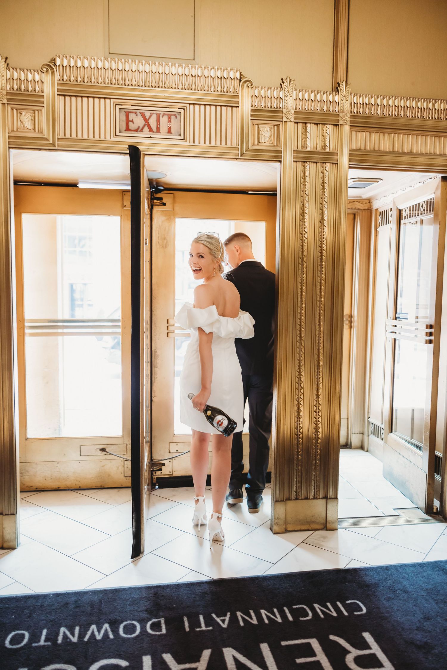 Bride in white mini dress holds champagne at Renaissance Cincinnati Downtown Hotel exit doors with groom.