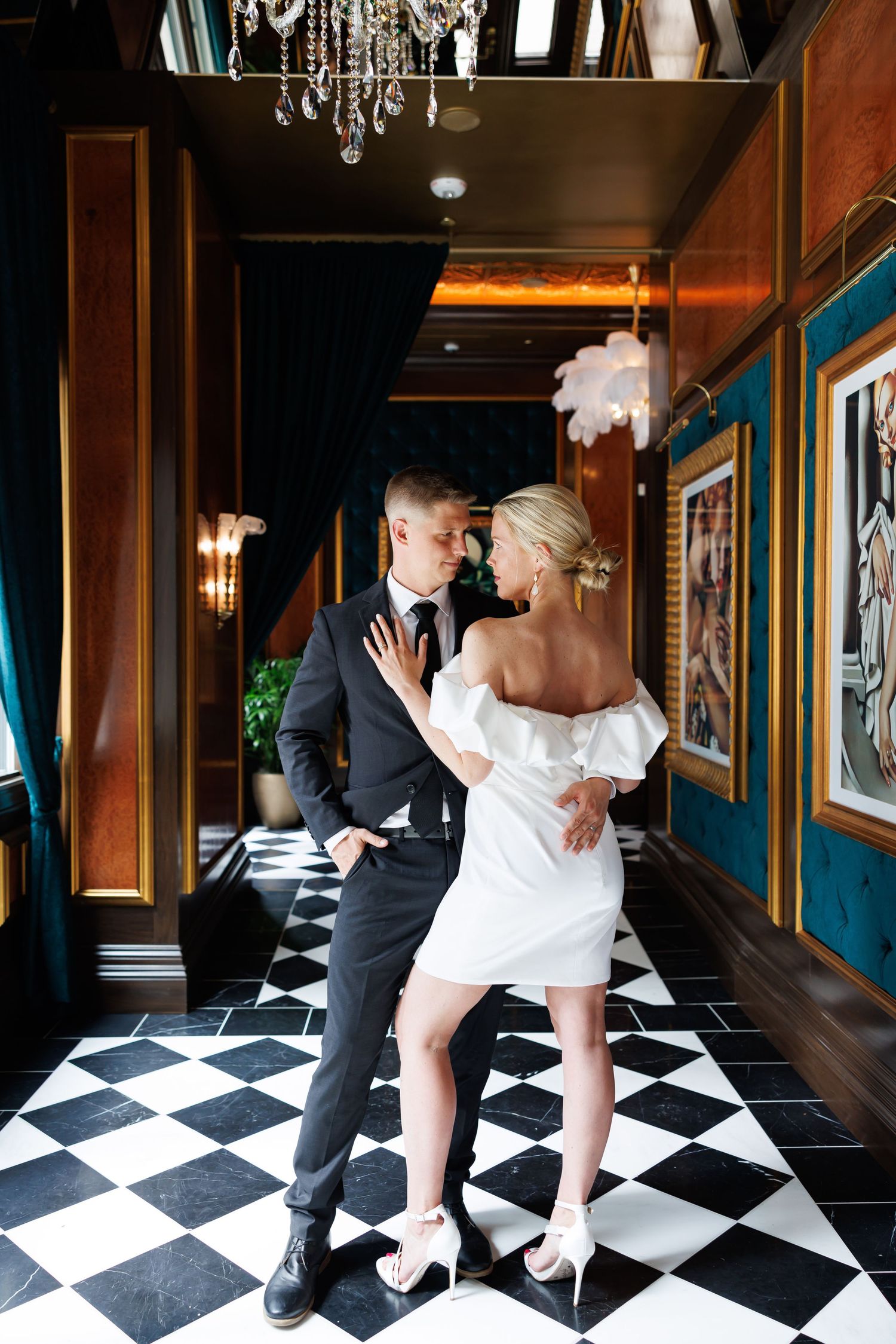 Elegant couple embracing in a luxurious hallway with checkered floors, crystal chandelier, and teal walls.