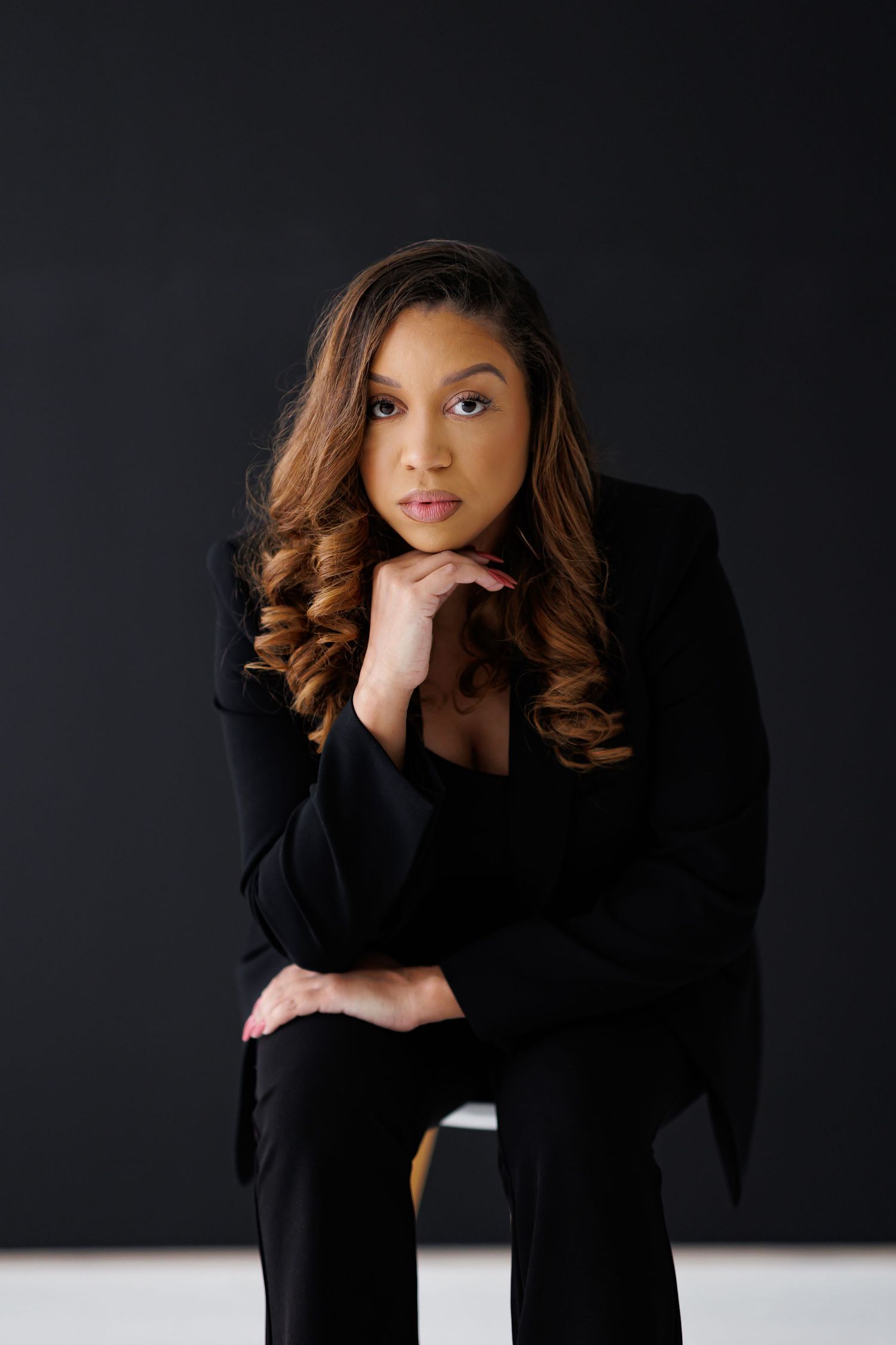Professional woman in black blazer and pants sits confidently against dark background with hands folded under chin.