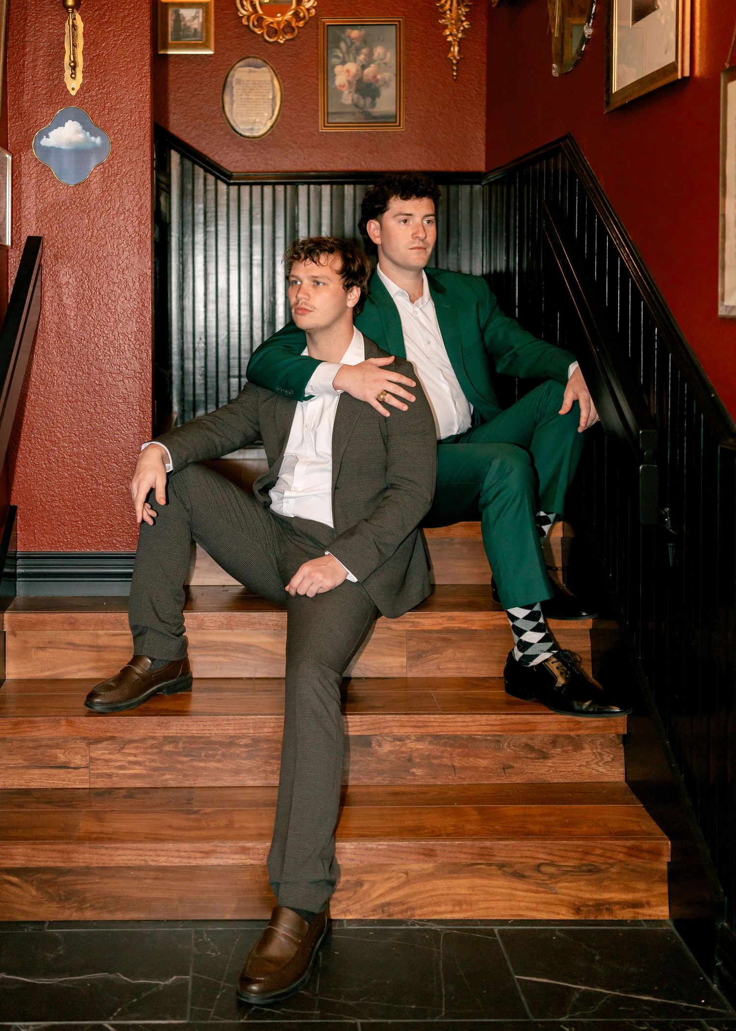 Two men in suits pose on wooden stairs in a red-walled interior, one in gray, one in green.