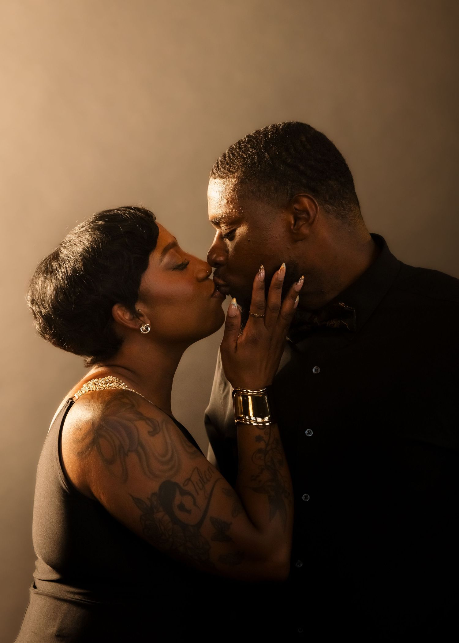 A couple sharing a romantic kiss, the woman with tattoos and short hair, the man in a black shirt, against a warm backdrop.