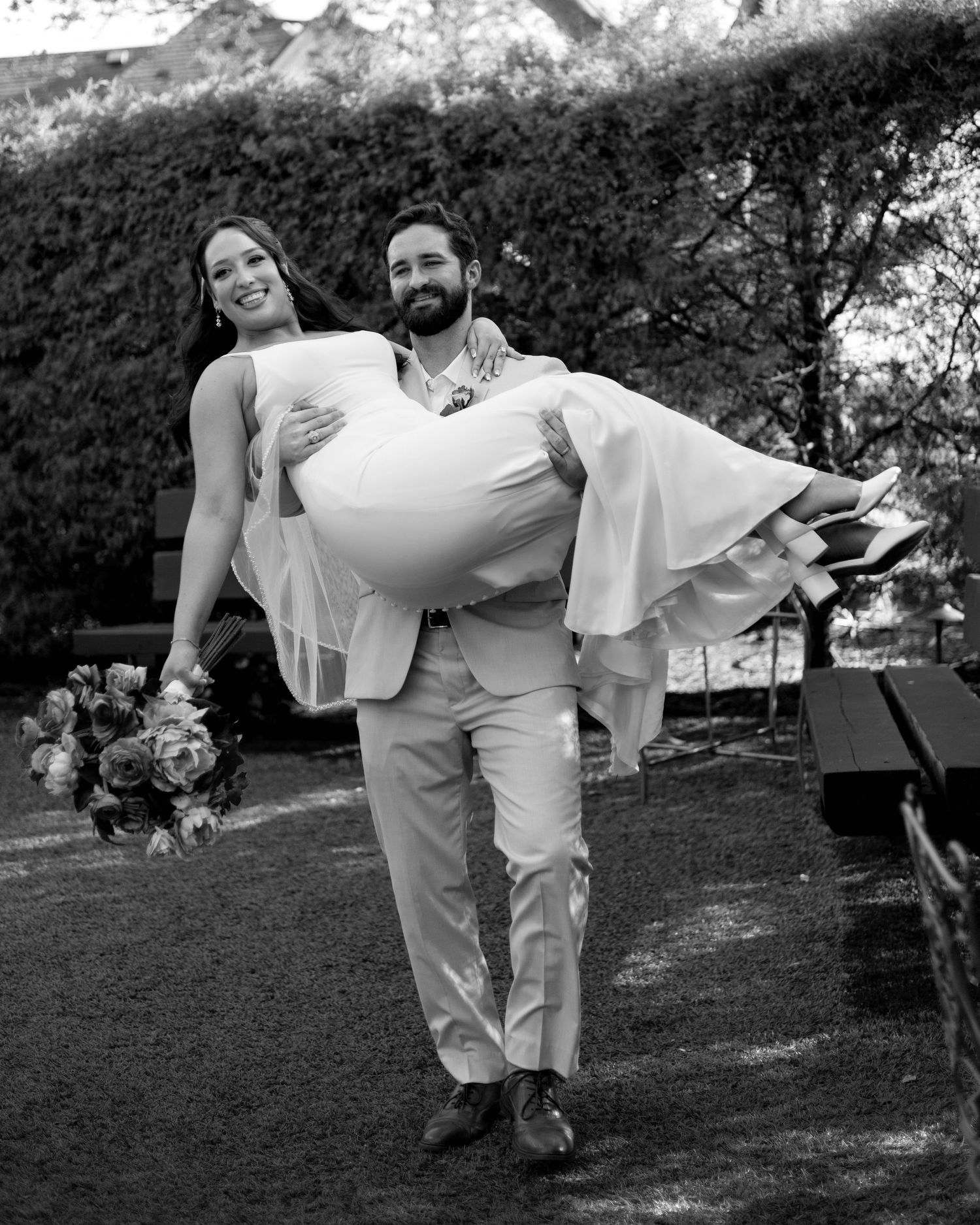 Black and white photo of a groom carrying his bride in a wedding dress outdoors, both smiling joyfully.