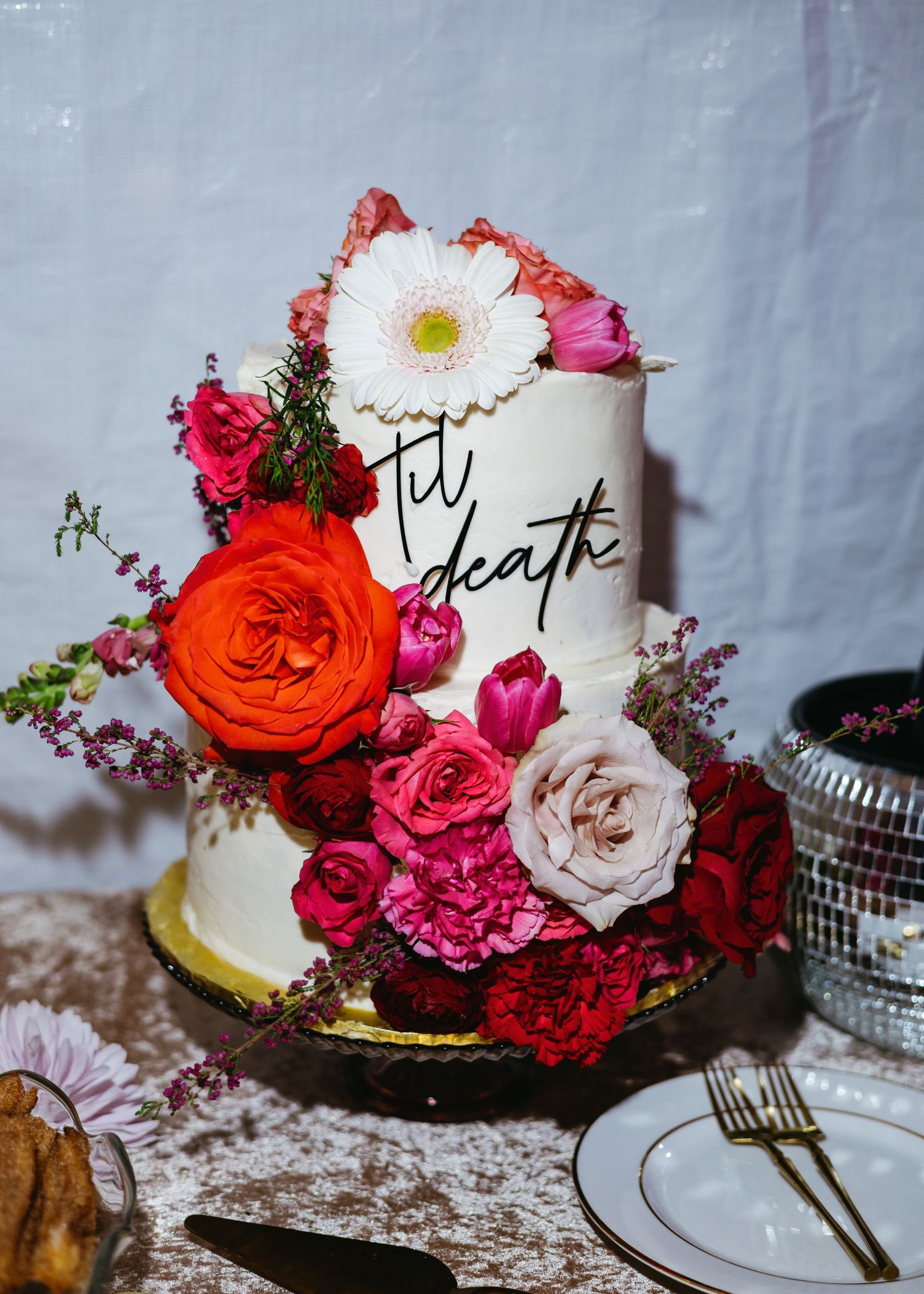 Two-tier white wedding cake adorned with vibrant red, pink, orange flowers and 'til death' calligraphy script decoration.