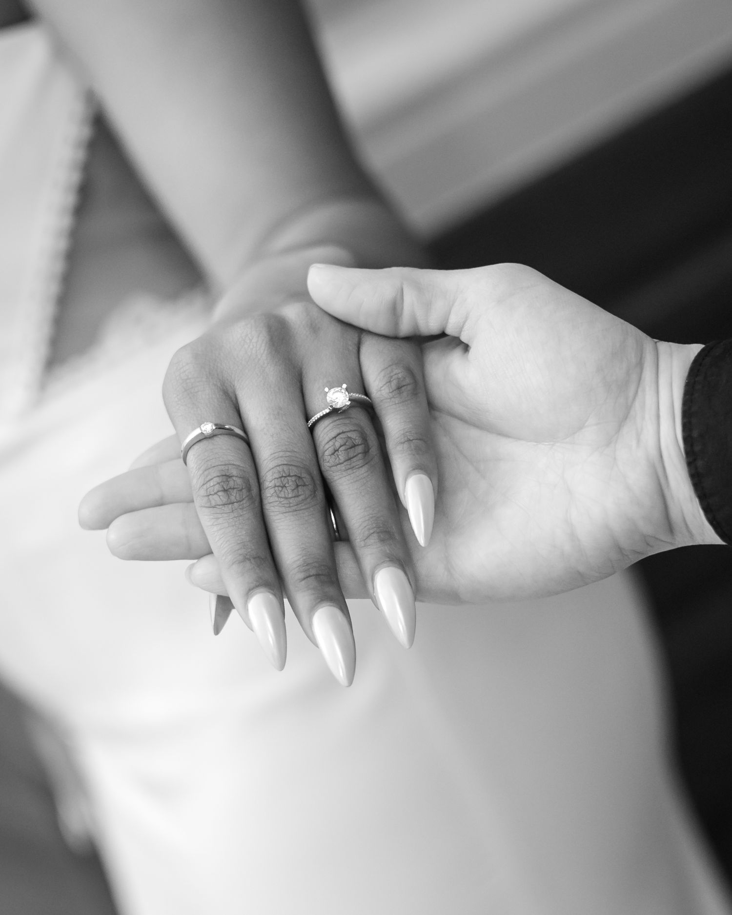 Black and white photo of a couple holding hands showing wedding rings and long white stiletto nails.